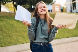 Pretty cheerful casual student girl opening envelope with exams results rejoicing in city park