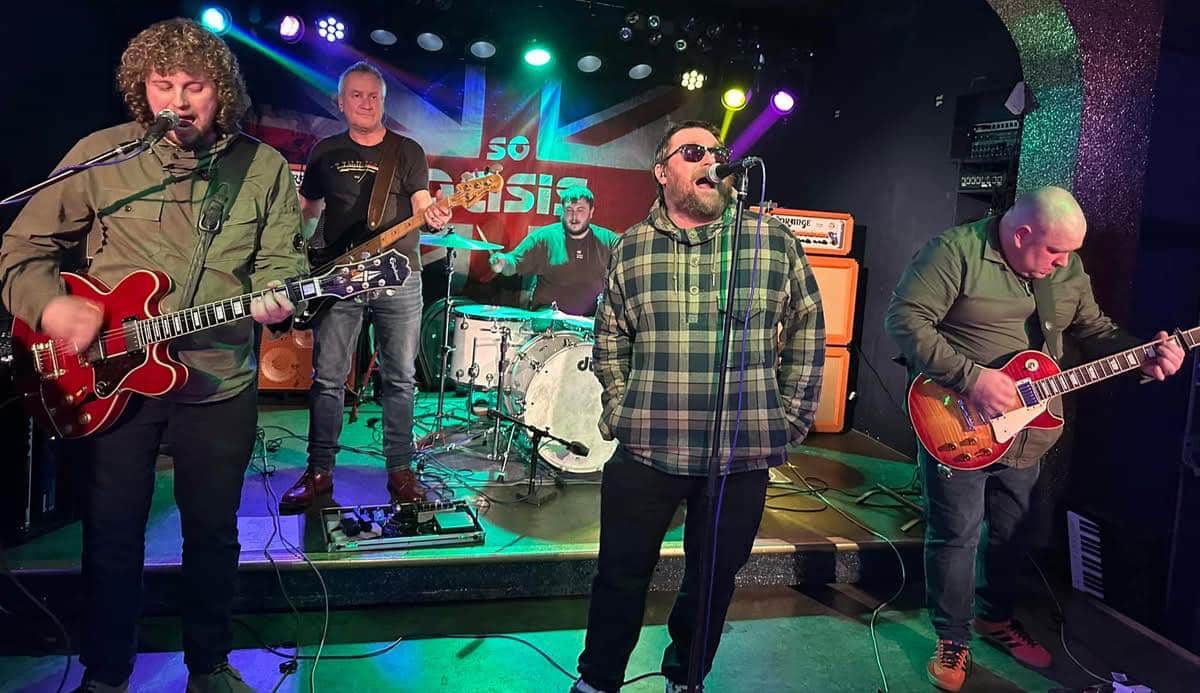 So Oasis tribute band performing live on stage with guitars, drums, and vocals, under colourful stage lights and a Union Jack backdrop.