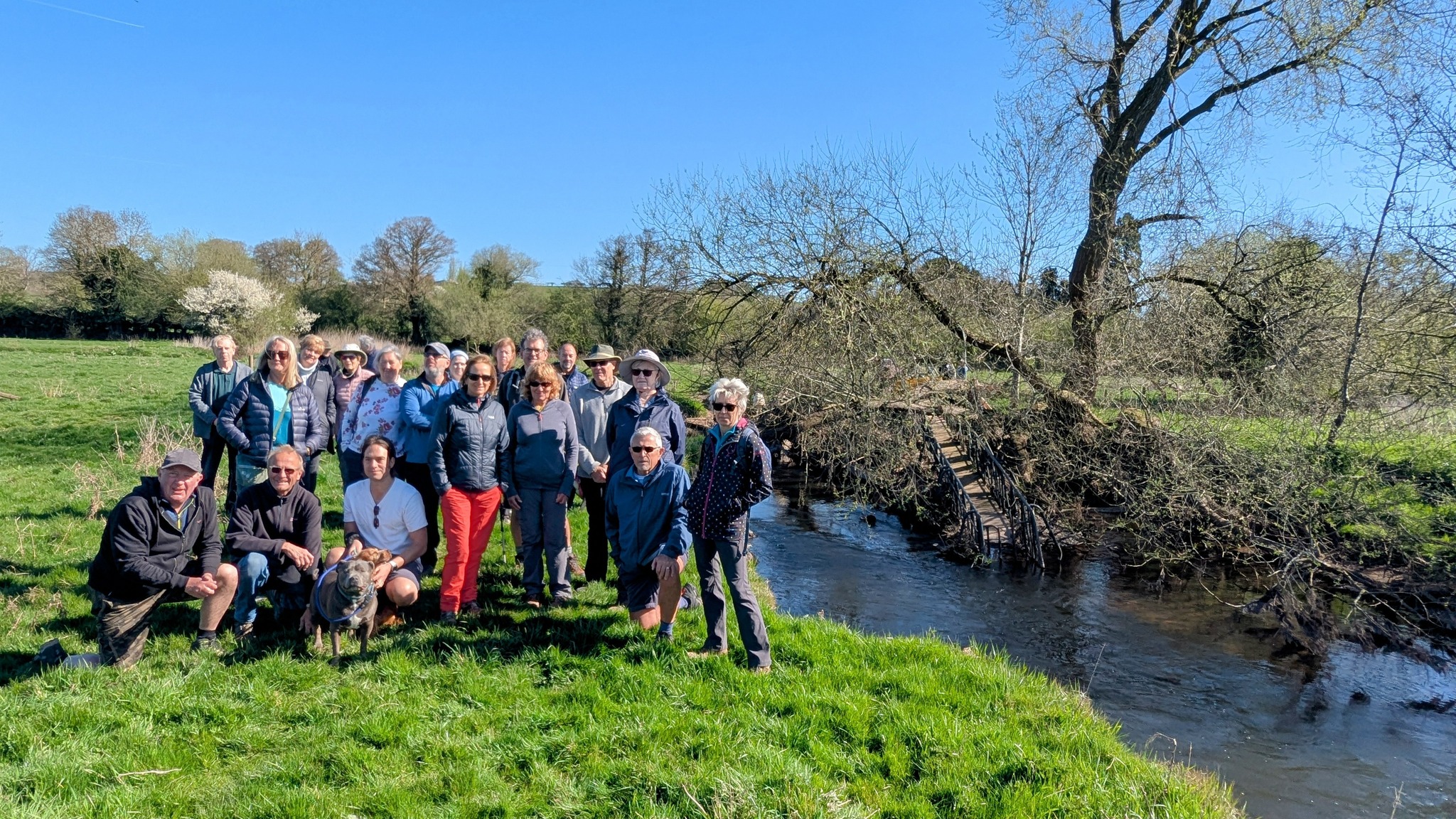 A group of around 20 local residents and a dog gathered on a grassy bank beside the damaged Aston to Burston footbridge, with clear blue skies and spring trees in the background. The broken wooden bridge is visible behind them, with fallen branches over the water.
