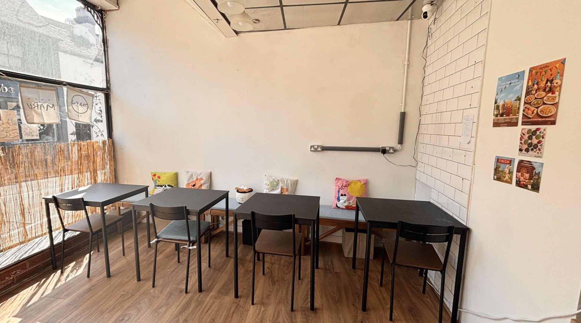 Black tables and chairs in Maru Bento. They sit on a wooden floor, with a cream wall behind and a window looking out to Radford Street in Stone.