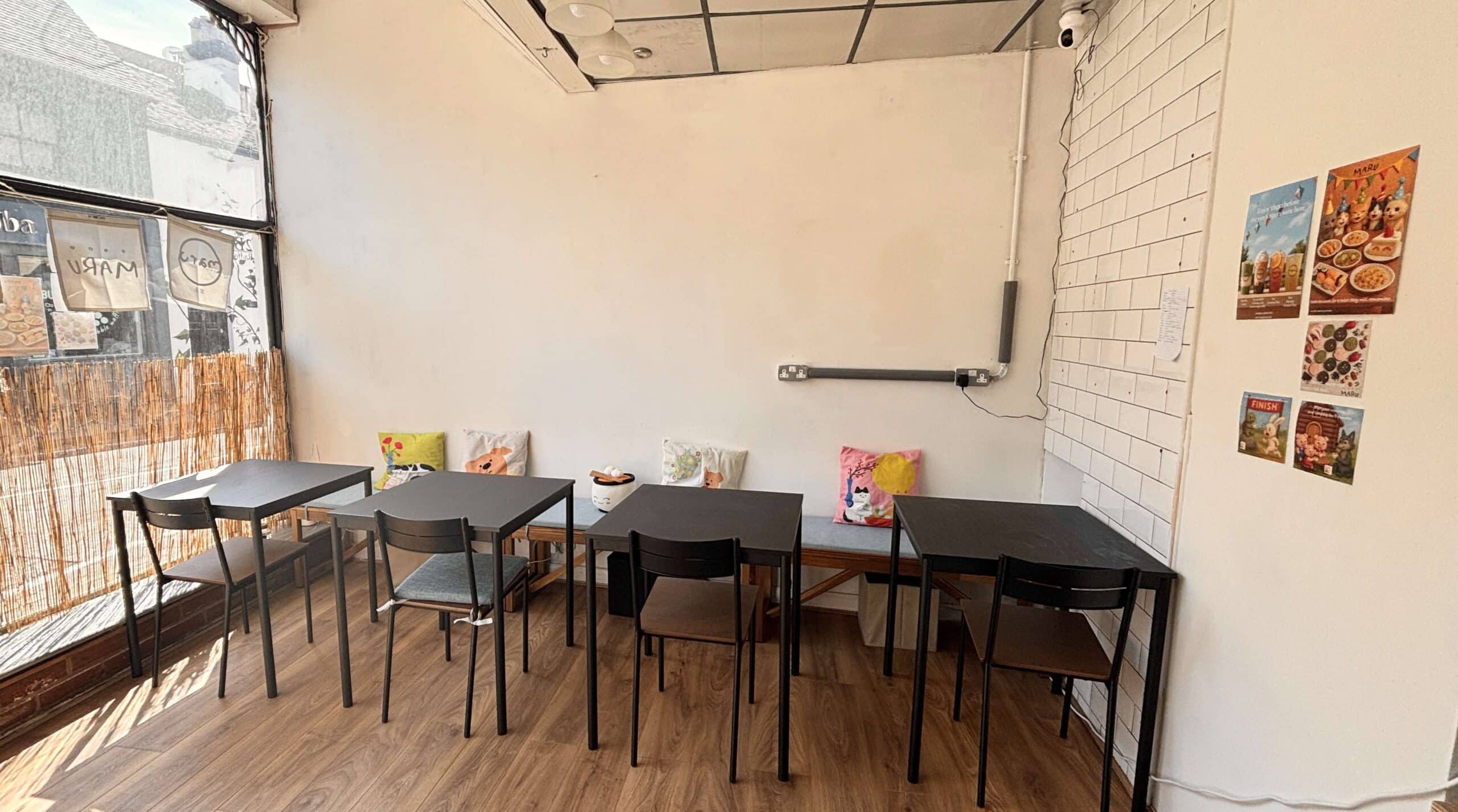 Black tables and chairs in Maru Bento. They sit on a wooden floor, with a cream wall behind and a window looking out to Radford Street in Stone.