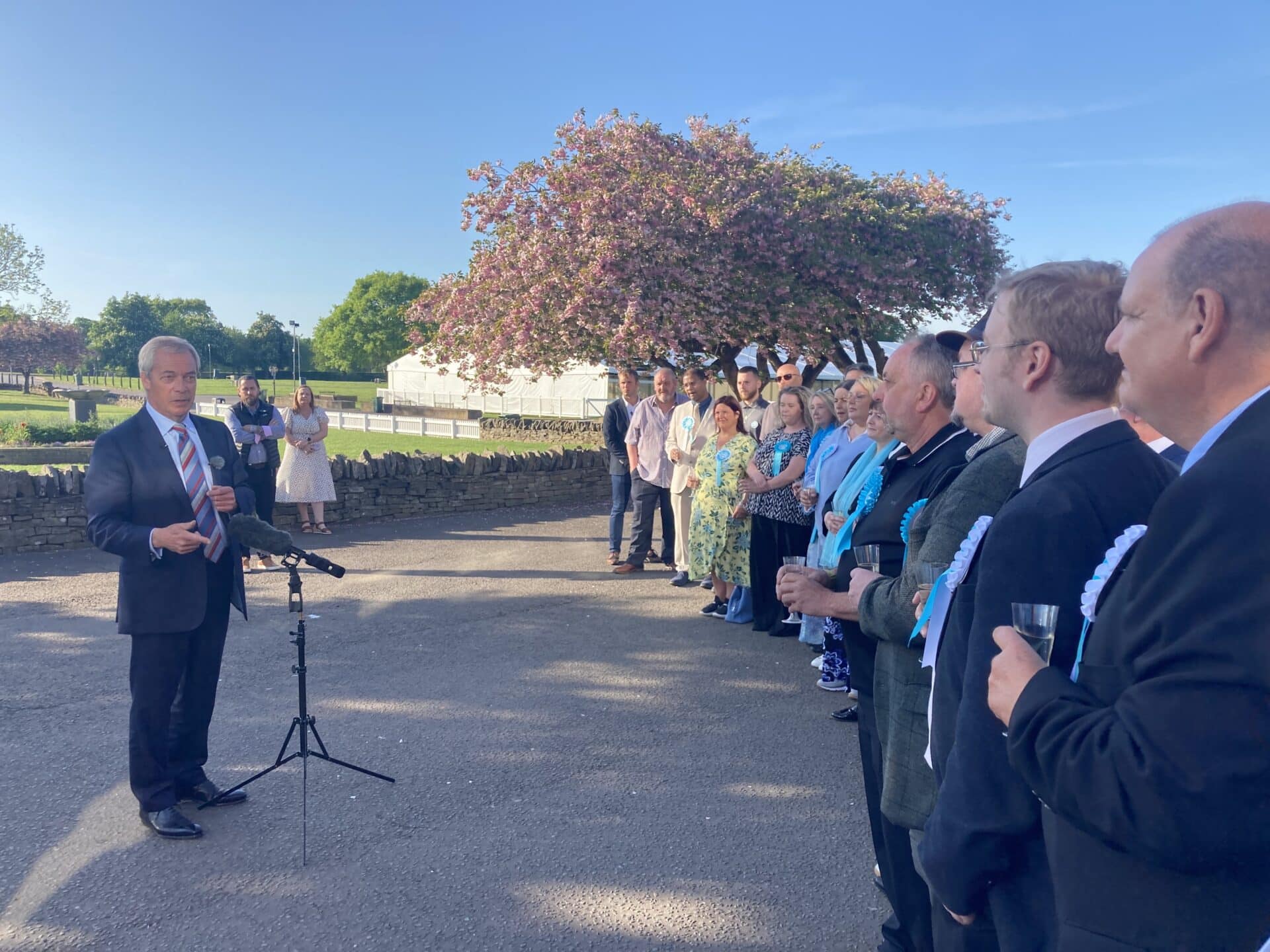 Nigel Farage addresses a line of newly elected Reform UK councillors and supporters during an outdoor speech at Stafford Showground, with cherry blossoms and a white marquee visible in the background.