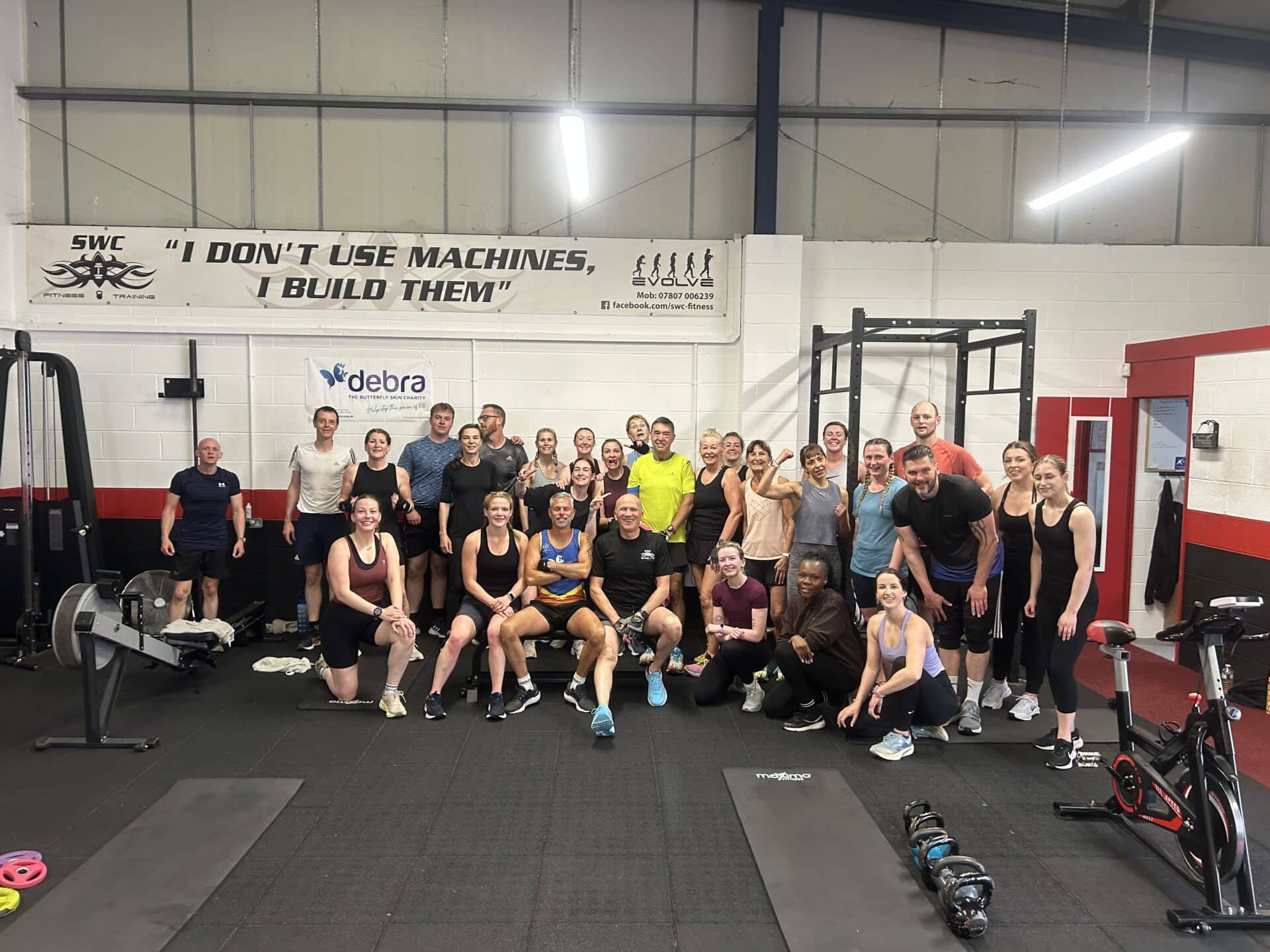 Group photo of SWC Fitness gym members and supporters gathered after a charity circuit session, with smiling participants posing in front of a DEBRA banner and gym equipment inside the Stone Business Park facility.