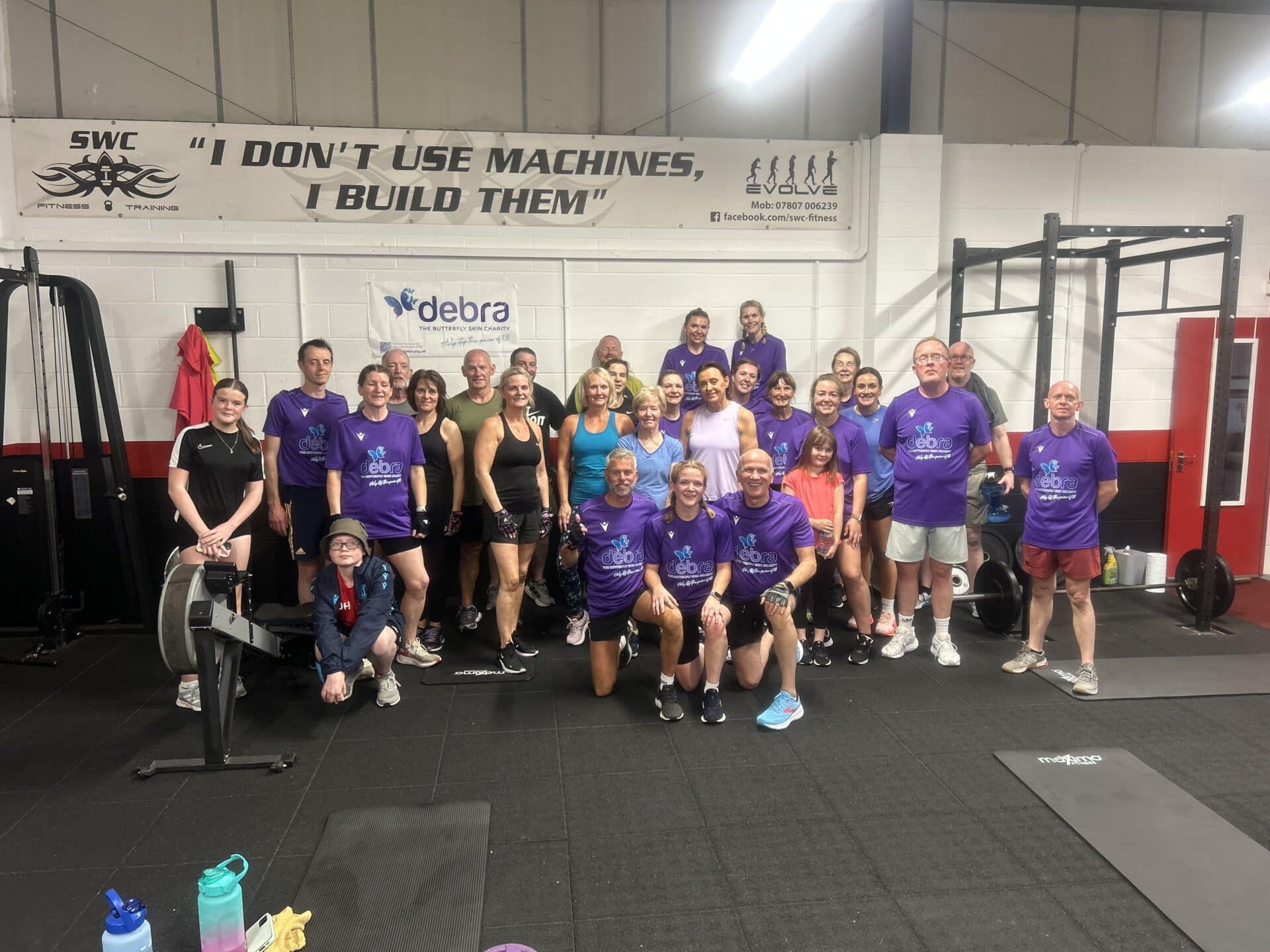 Group of SWC Fitness members and supporters wearing purple DEBRA charity T-shirts posing after a fundraising circuit session, smiling together in the gym at Stone Business Park beneath a banner reading “I don’t use machines, I build them.”