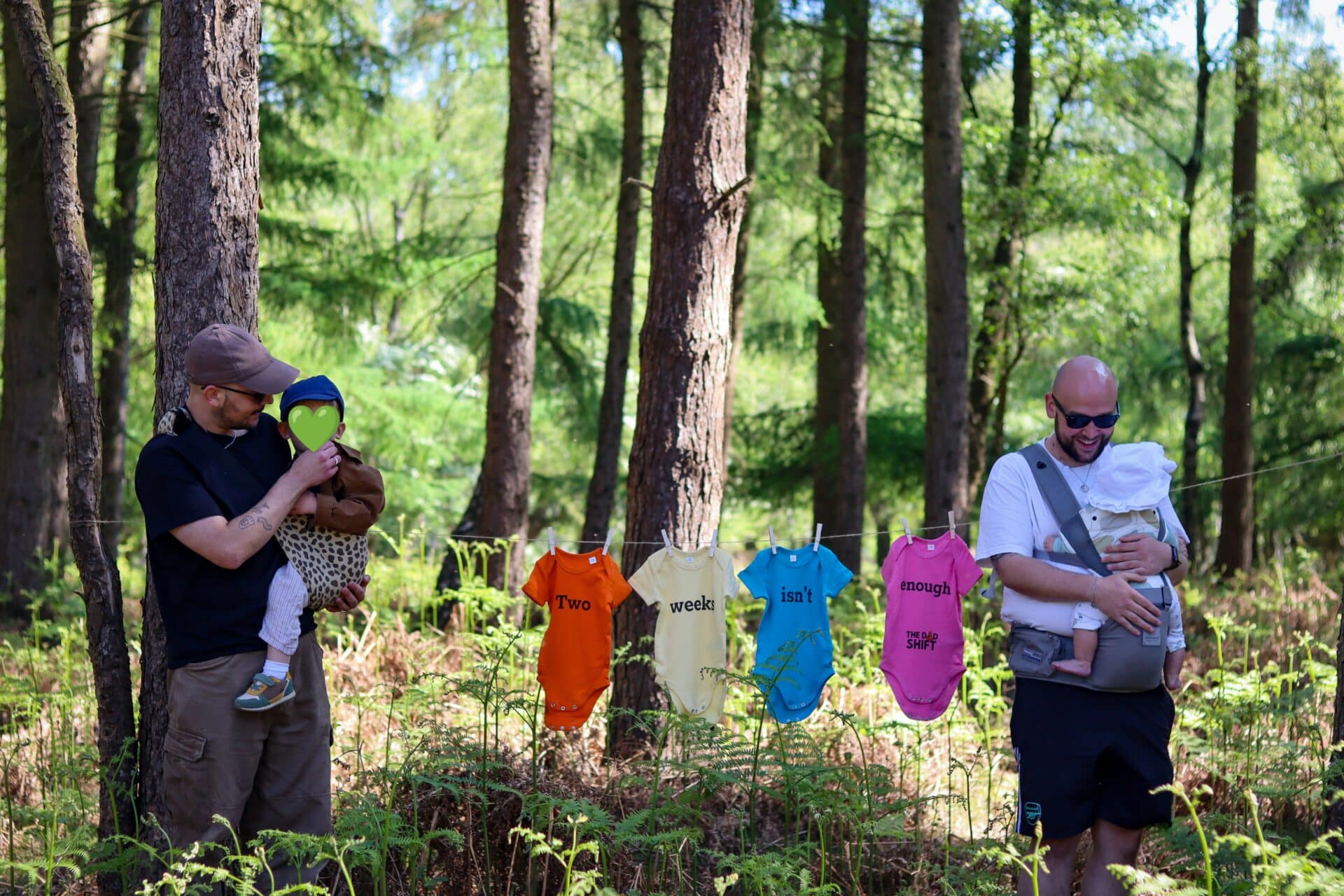 Two Stone dads stand in Cannock Chase Forest with their babies. In between them, baby grows on a washing line spell out the message “Two weeks isn’t enough” in support of better paternity leave.