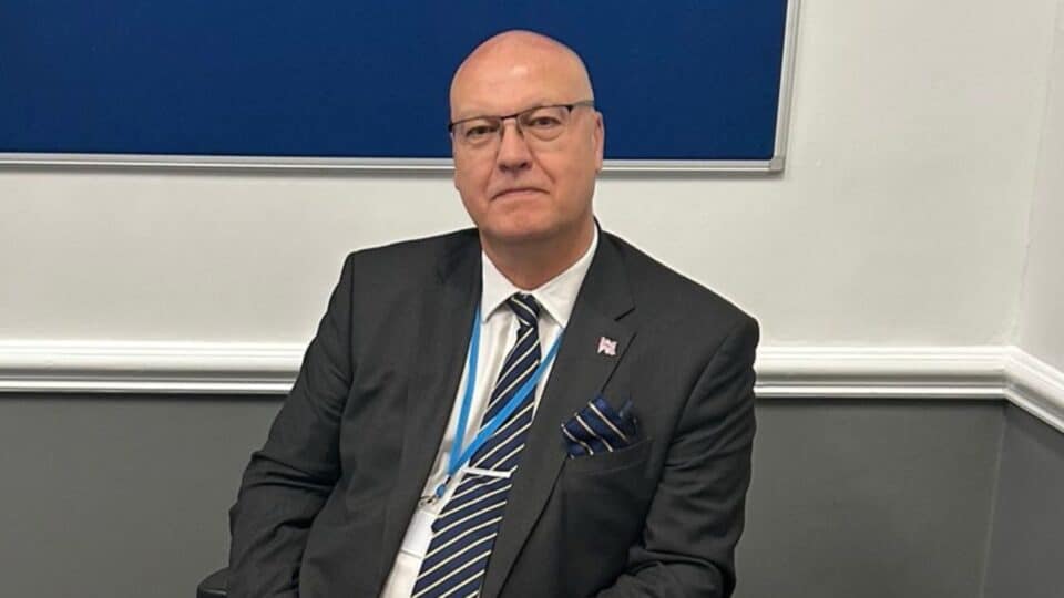 Ian Cooper, dressed in a dark suit and striped tie, sits against a two-tone wall with a blue noticeboard behind him. He wears glasses and a small Union Jack pin on his lapel.