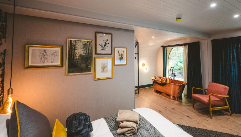 Interior of a Moddershall Oaks woodland pod featuring a copper roll-top bath, mid-century armchair, and a gallery wall of nature-inspired artwork above a plush king-size bed.