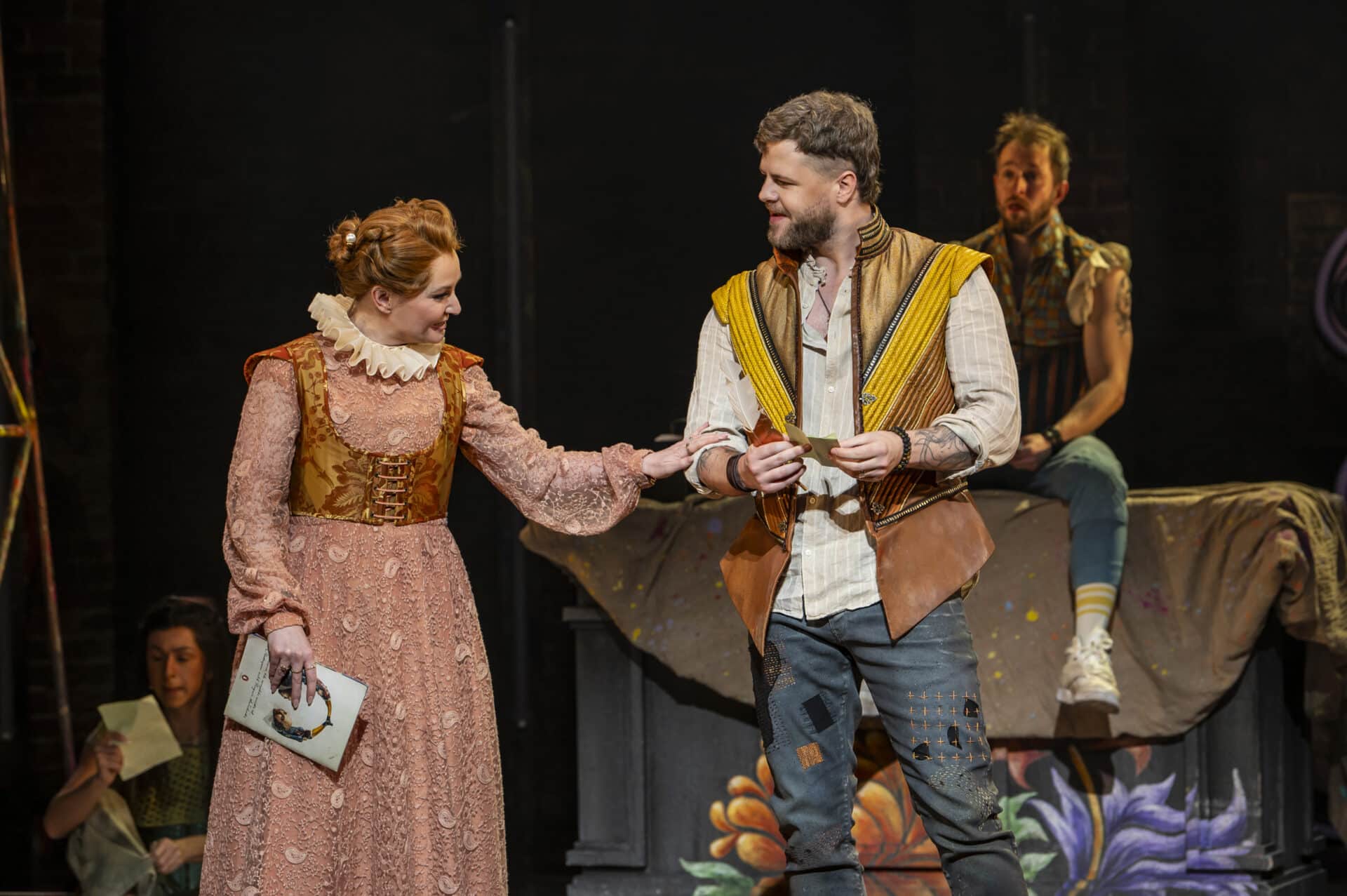 Lara Denning as Anne Hathaway and Jay McGuiness as Shakespeare share a moment on stage in & Juliet, with Anne holding a script and both dressed in Elizabethan-inspired costumes, lit warmly against a theatrical backdrop