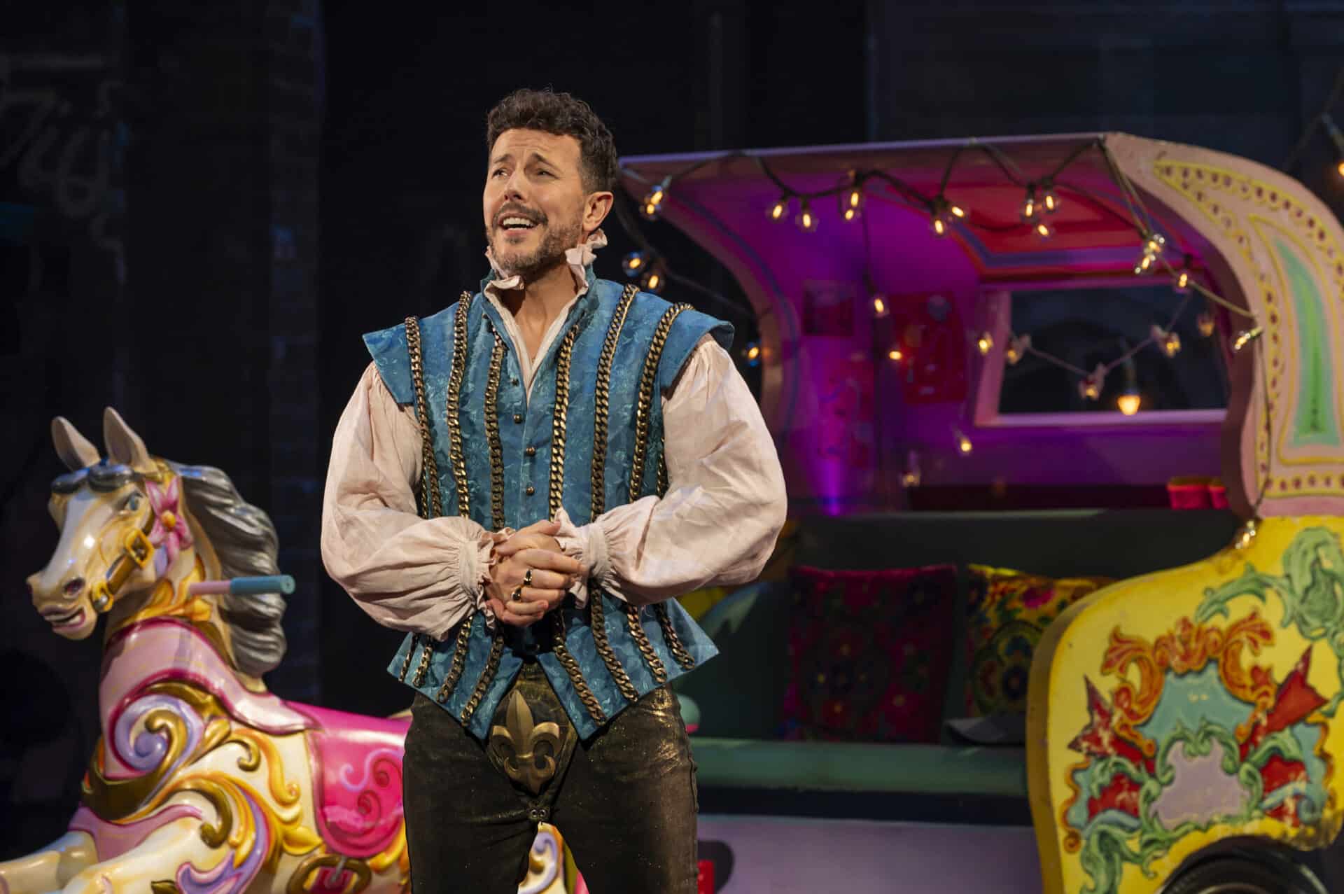 Lee Latchford-Evans as Lance in a richly embroidered blue waistcoat stands beside a brightly coloured fairground carousel horse and wagon, mid-performance in & Juliet
