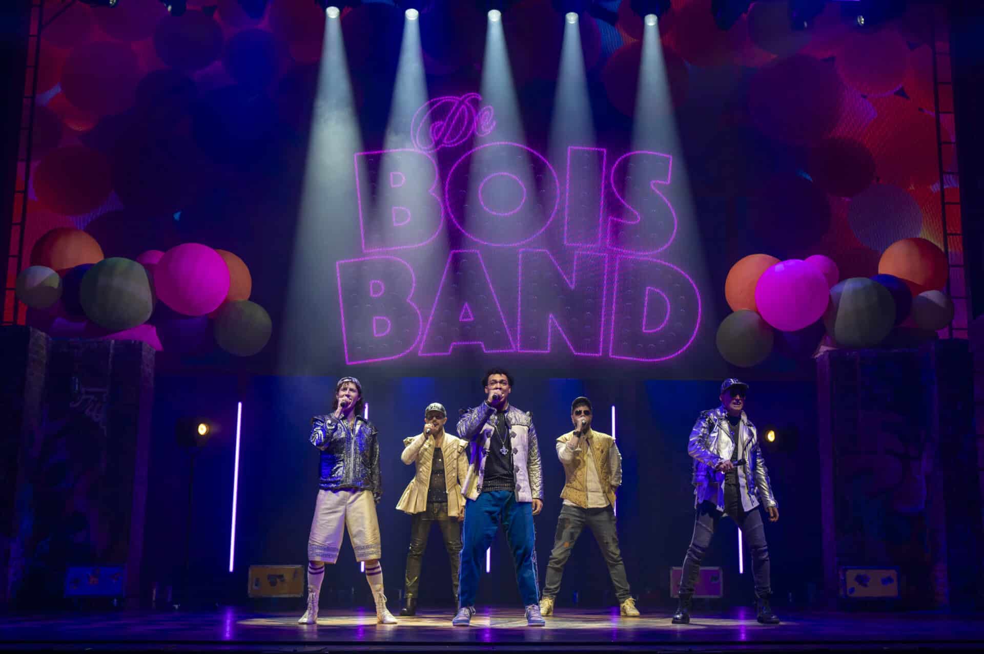 The De Bois Band characters perform under bright stage lights with a glowing pink backdrop, dressed in glittery and urban outfits in a lively boyband parody scene from & Juliet