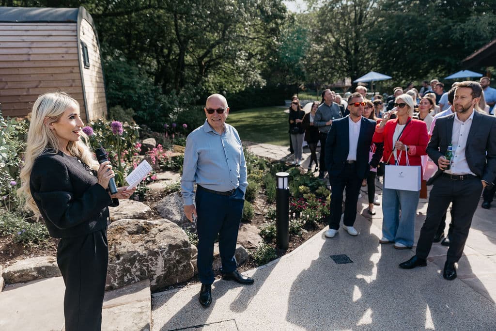 Moddershall Oaks director Penny Weston addresses guests with a microphone during the official launch event for the new woodland pods, with a pod and floral landscaping visible in the background.