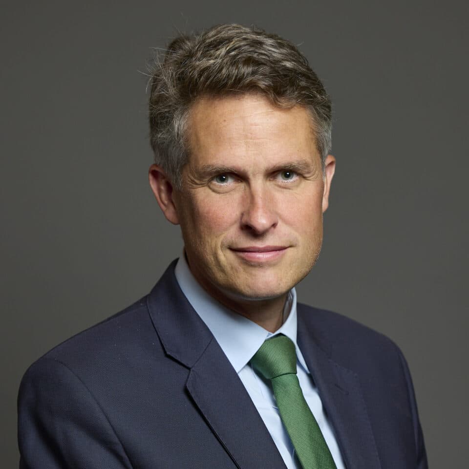 Official portrait of Sir Gavin Williamson, MP for Stone, Great Wyrley and Penkridge, wearing a navy blue suit, light blue shirt, and green tie, set against a plain grey background.