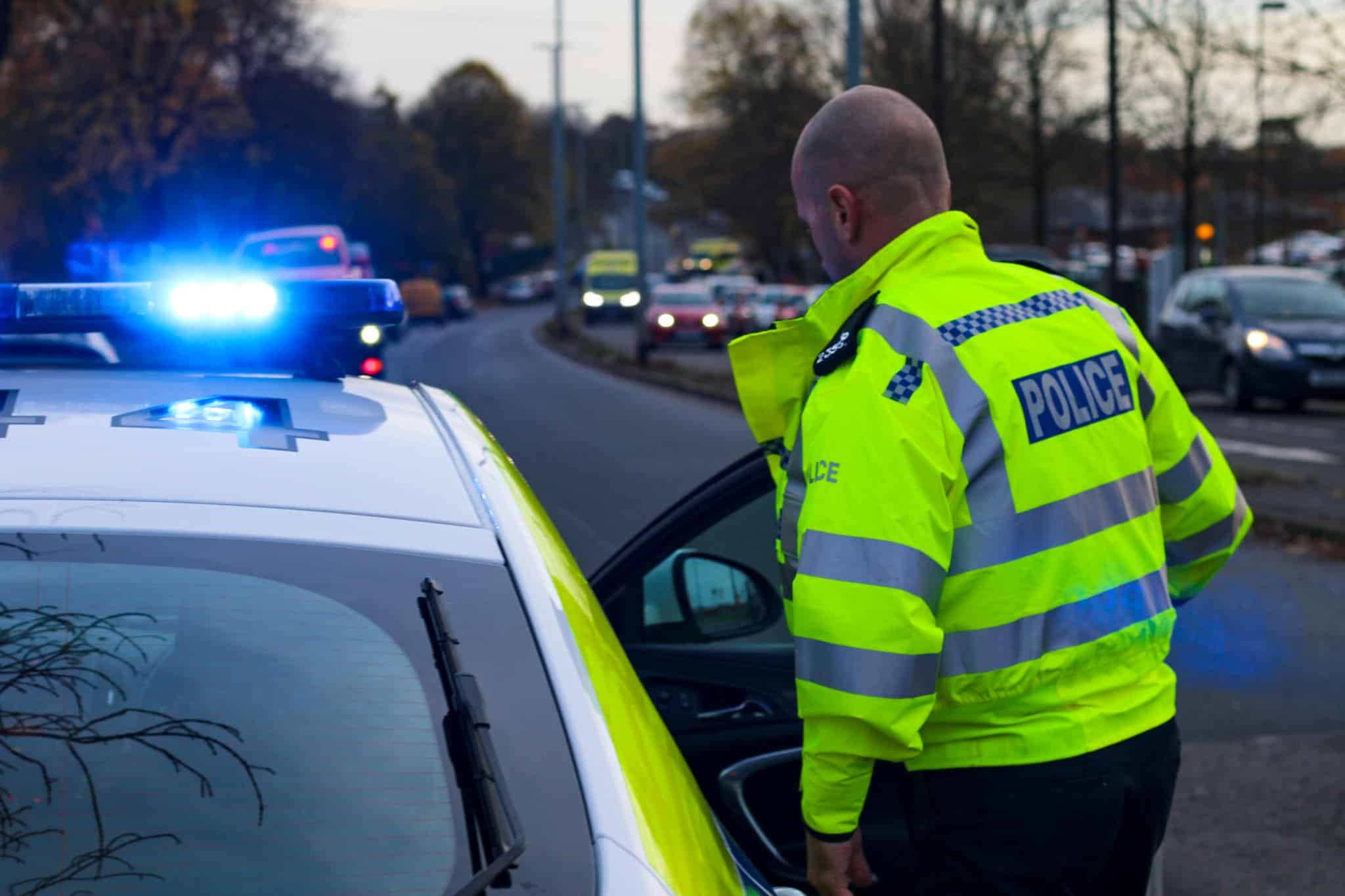 A Staffordshire Police officer in a high-visibility jacket stands next to a marked police car with blue lights flashing. The car is parked at the side of a busy road during daylight, with other vehicles and trees in the background.
