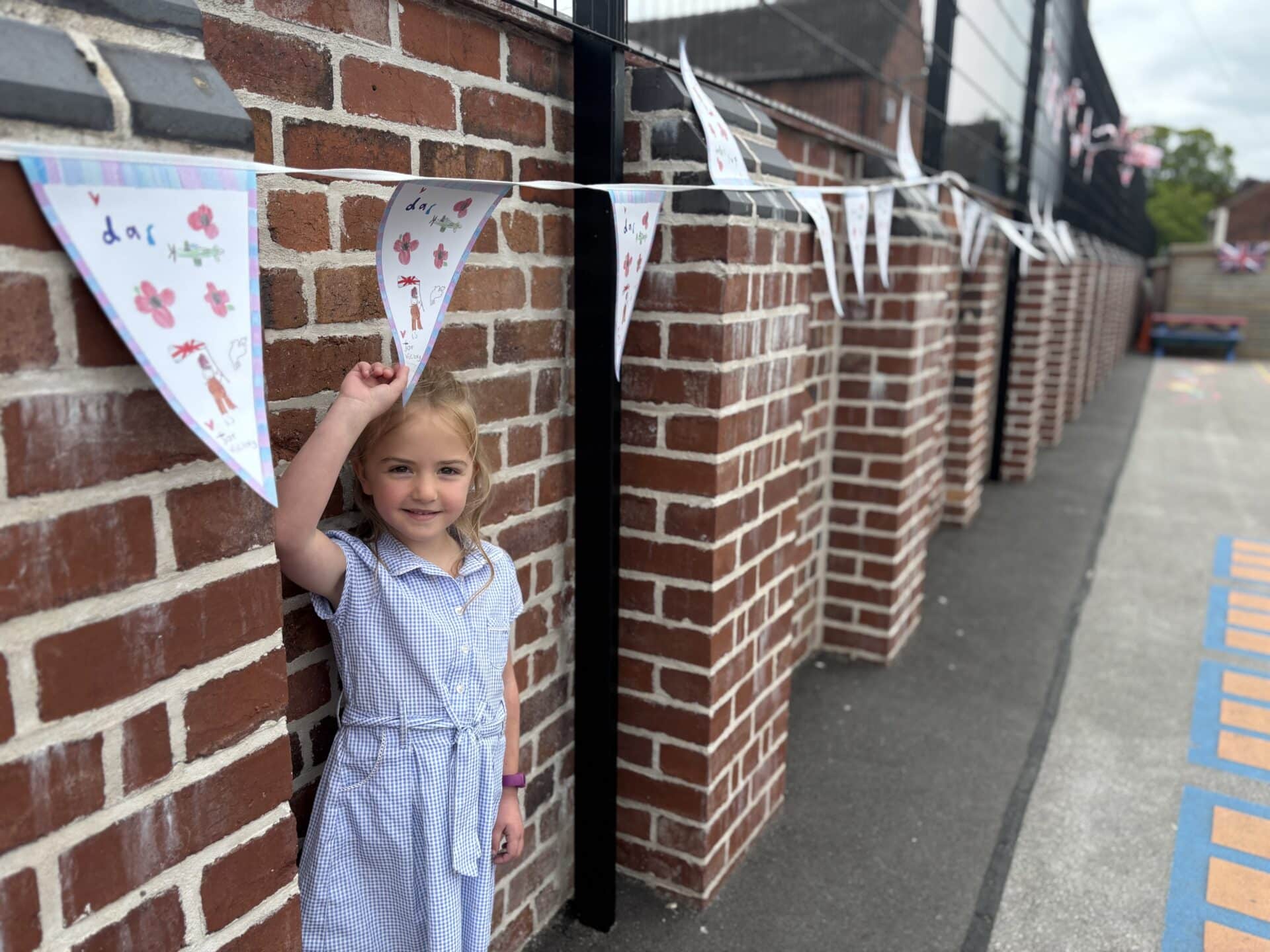 Lauren Goodman, winner of the Stone VE Day bunting competition, smiles in her school playground as she points to her winning bunting displayed along the brick wall.