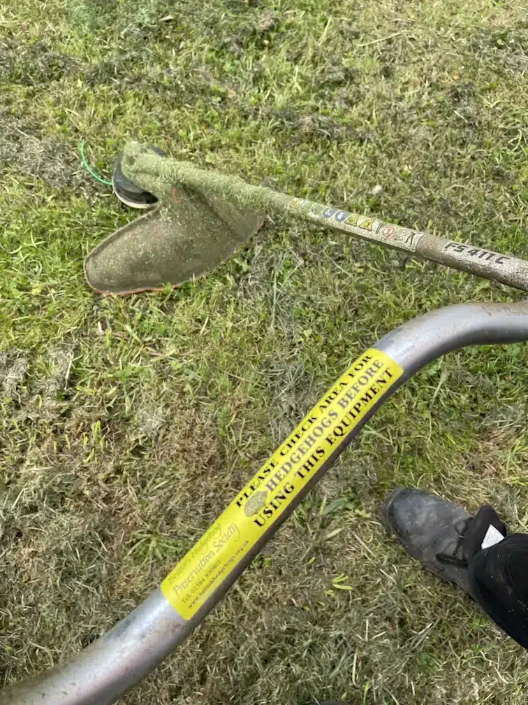 A council worker's strimmer featuring a bright yellow warning sticker from the British Hedgehog Preservation Society, reminding users to check for hedgehogs before use.