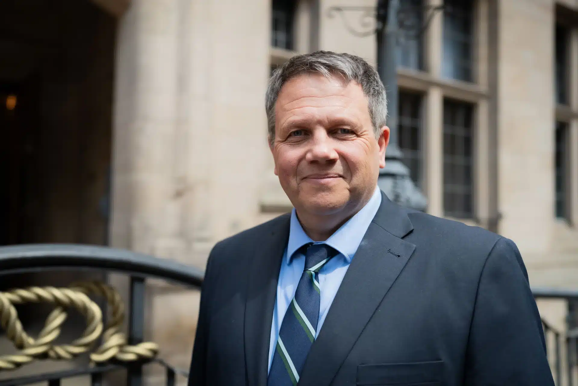 Andrew Mynors, Staffordshire County Council’s Cabinet Member for Connectivity, standing outside a civic building in a dark suit, light blue shirt, and striped tie, smiling at the camera.