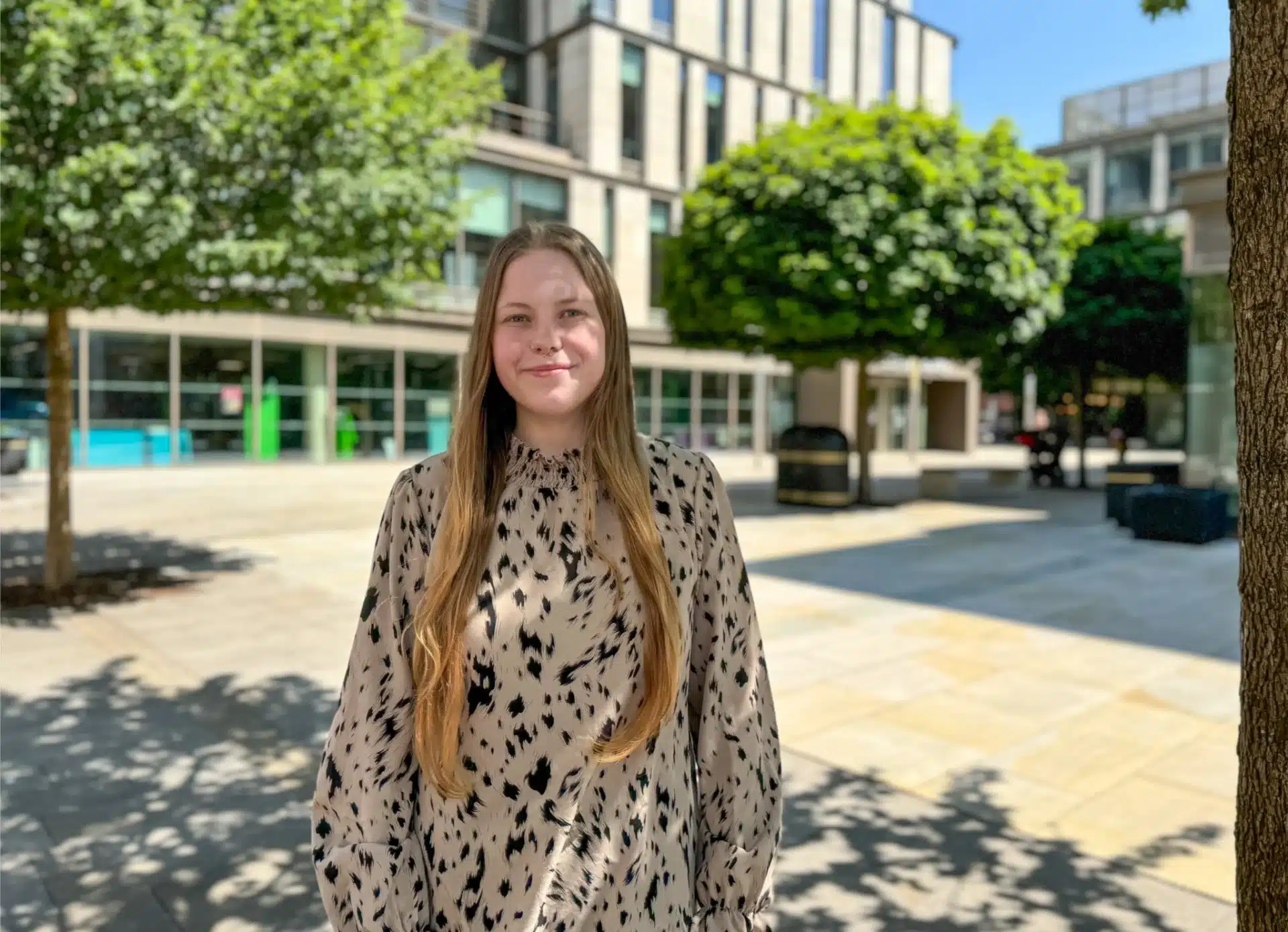 Molly Wynn, a young woman with long straight hair, wearing a patterned blouse, standing outdoors in a sunlit modern plaza with trees and office buildings in the background.