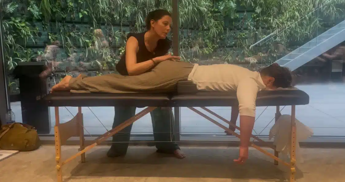 Sam Macpherson performing a Spinal Flow session, gently applying touch to a client lying face down on a therapy table in a calm, modern setting with a lush green plant wall in the background.