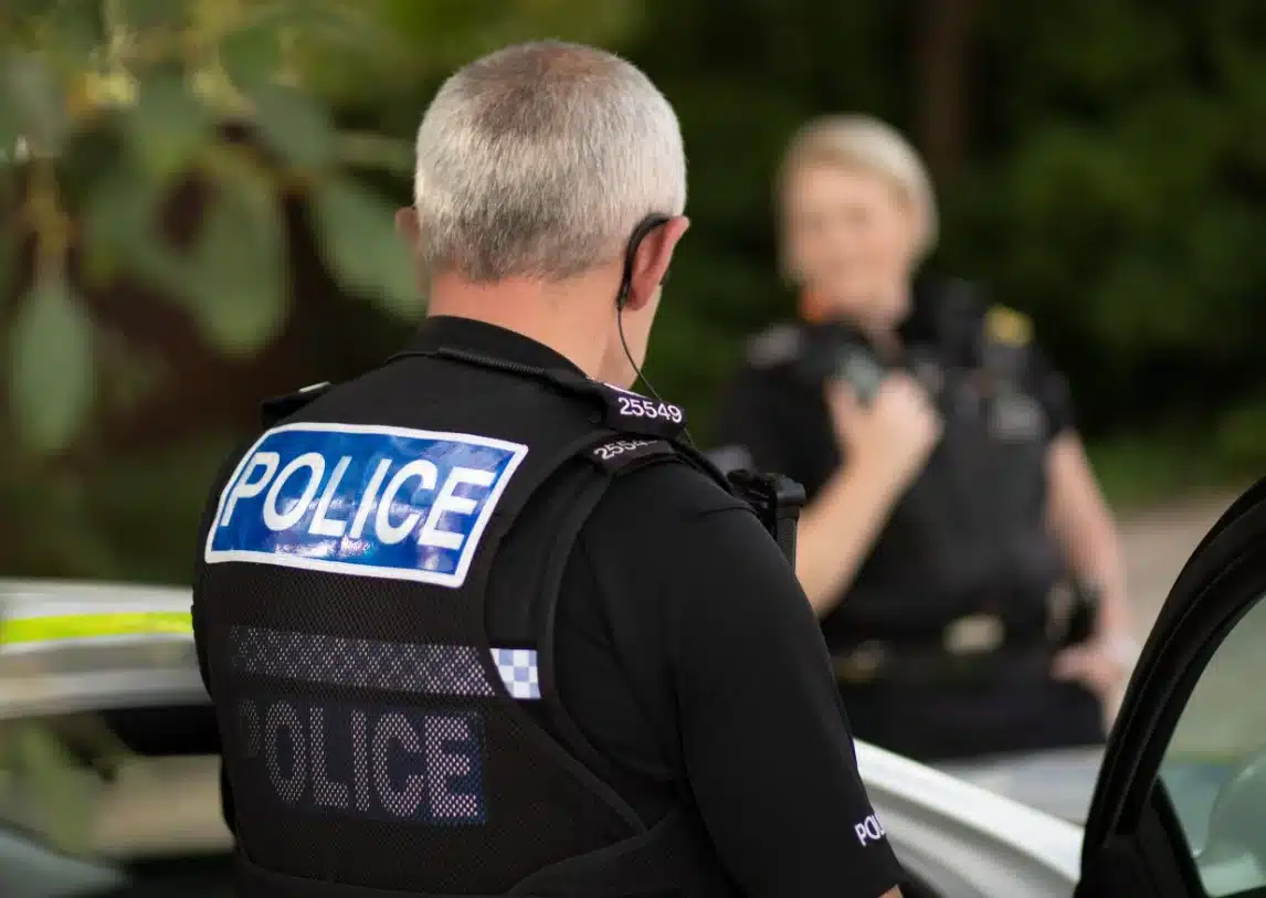 Two Staffordshire Police officers in uniform, one facing away wearing a vest marked "POLICE", and the other slightly out of focus in the background beside a patrol car.