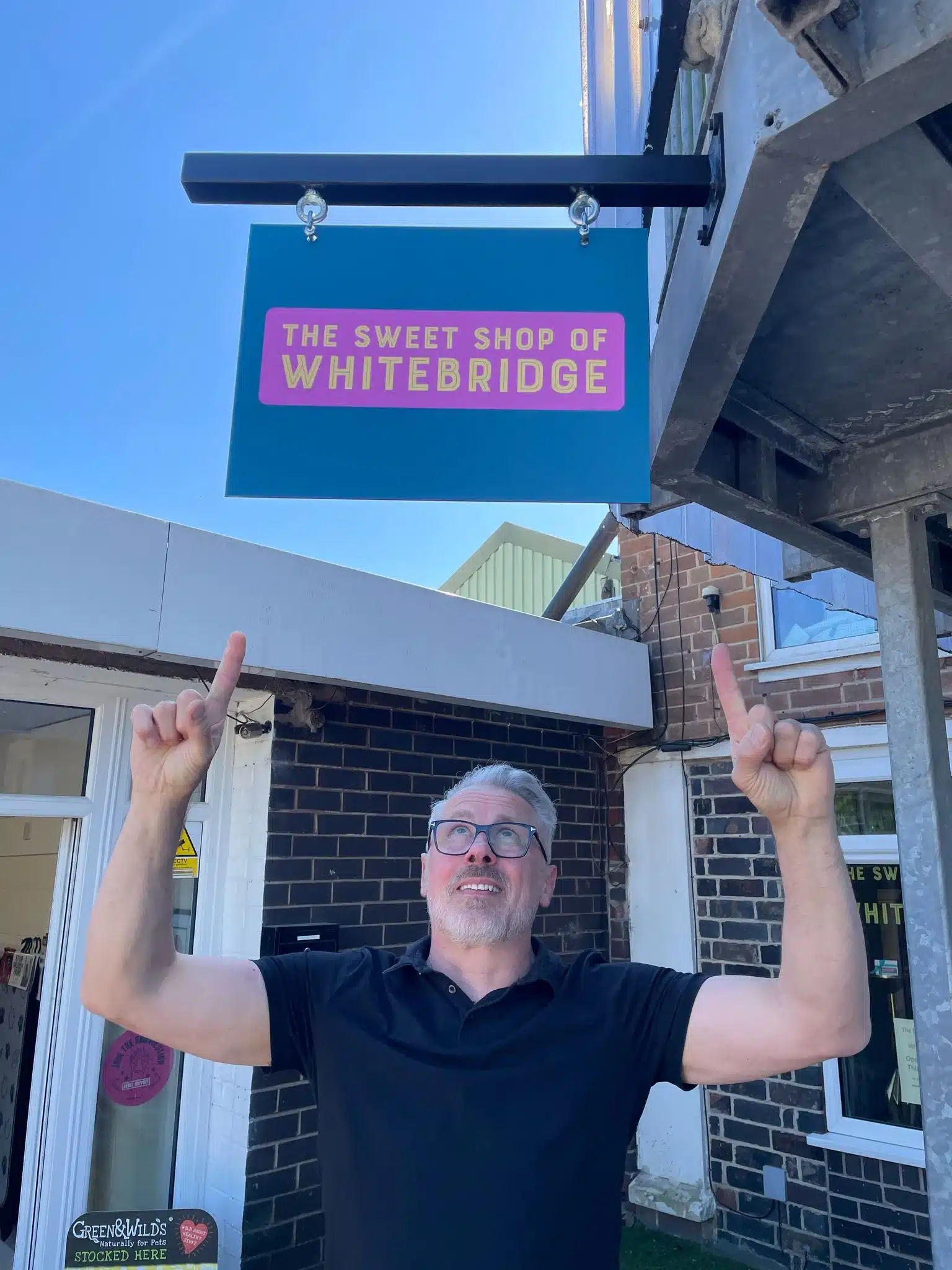 David Wolstenholme smiling and pointing proudly at the new hanging sign for The Sweet Shop of Whitebridge on Whitebridge Industrial Estate.