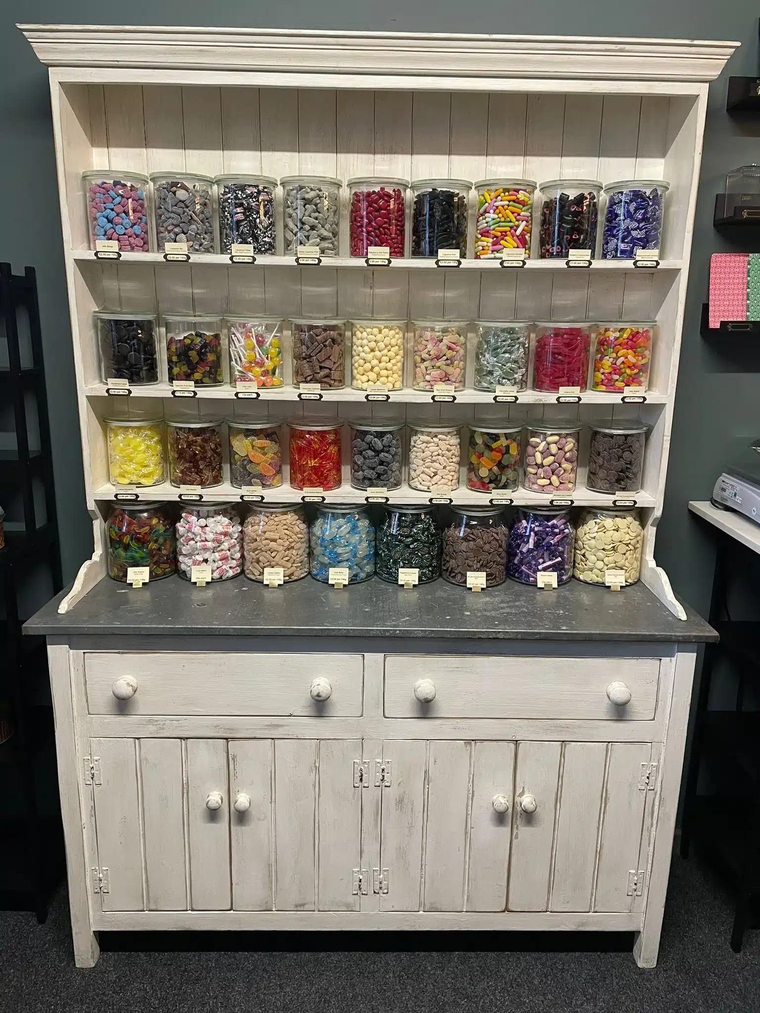 Rows of retro sweets and liquorice varieties in glass jars displayed on a white wooden dresser inside The Sweet Shop of Whitebridge.