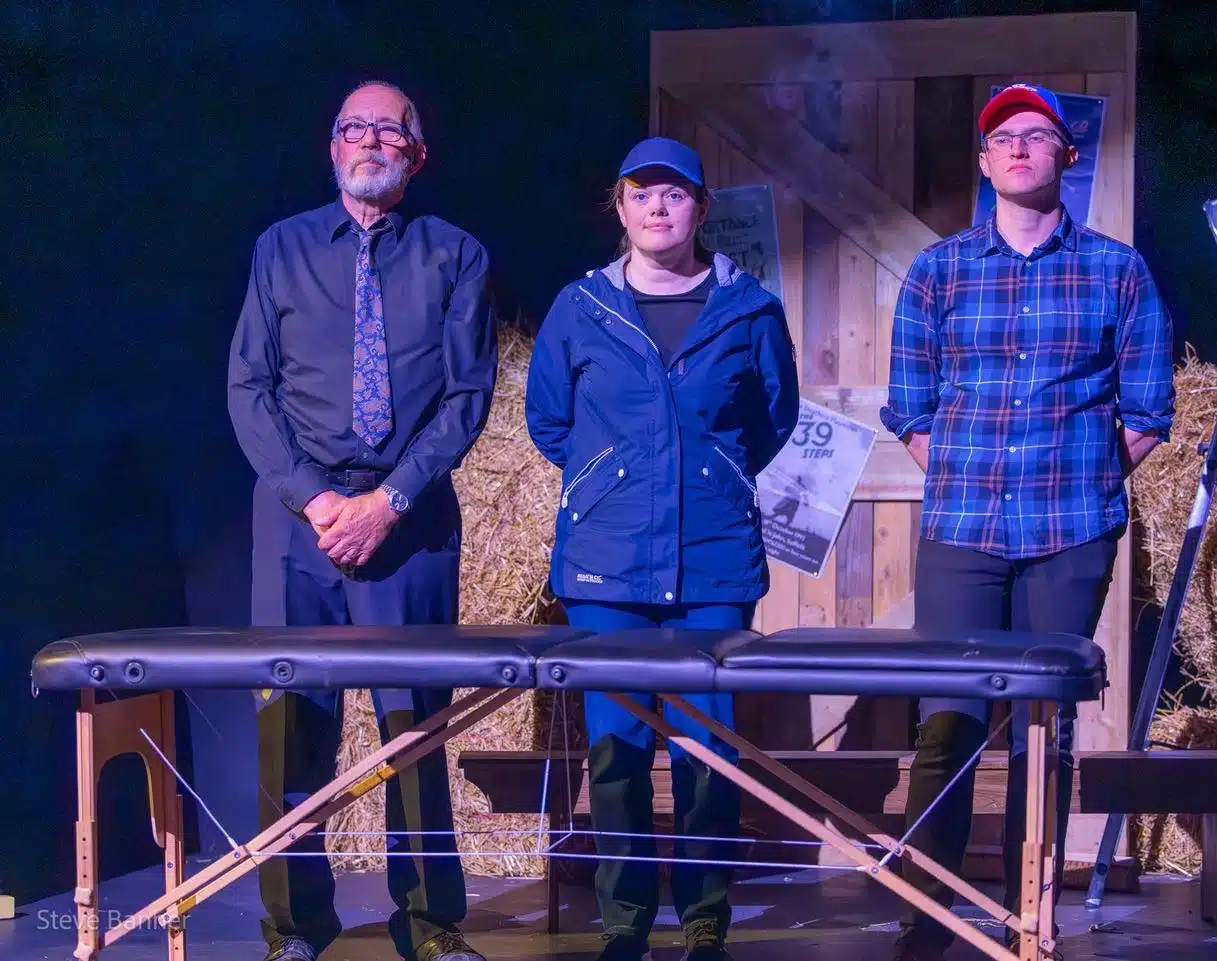 Three cast members from A Bunch of Amateurs stand on stage behind a massage table, framed by hay bales and rustic props at Crown Wharf Theatre.