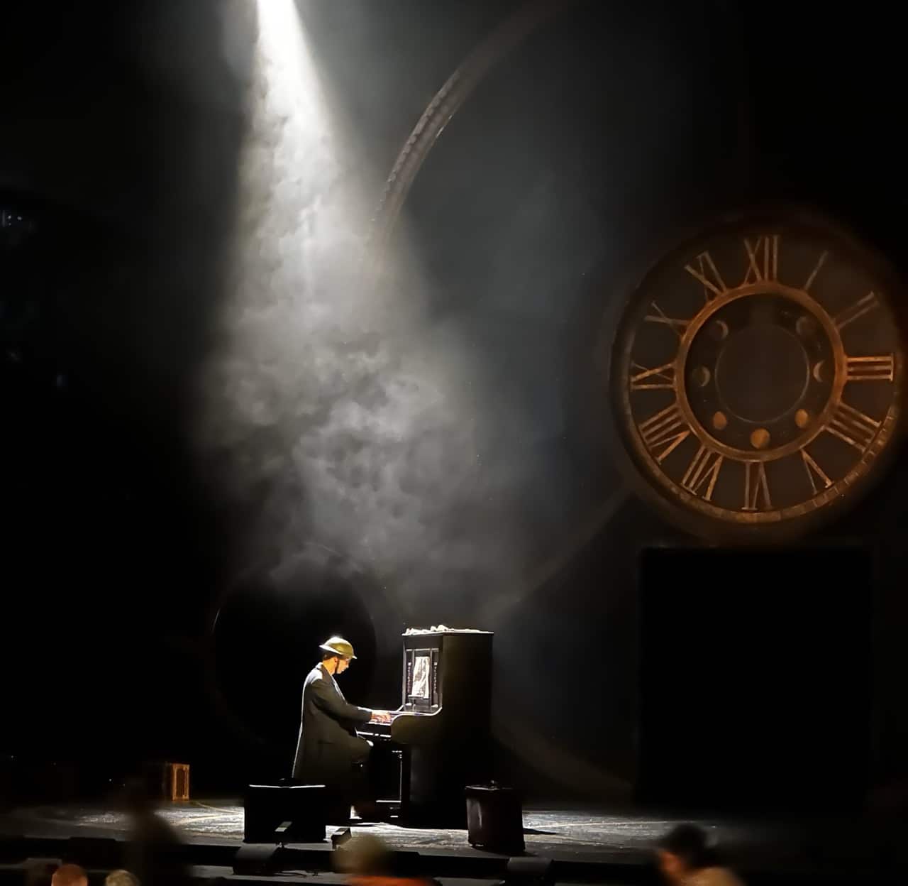 A spotlight shines down on a wartime soldier in uniform playing a piano on stage, with smoke swirling around and a large clock face looming in the background. The scene evokes a sense of nostalgia and sets the tone for the theatrical performance of The Lion, The Witch and The Wardrobe.