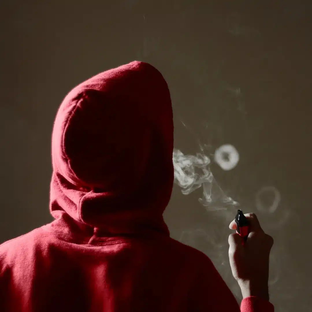 Teenager in a red hoodie using a vape, with visible smoke rings, highlighting concerns around underage vaping.