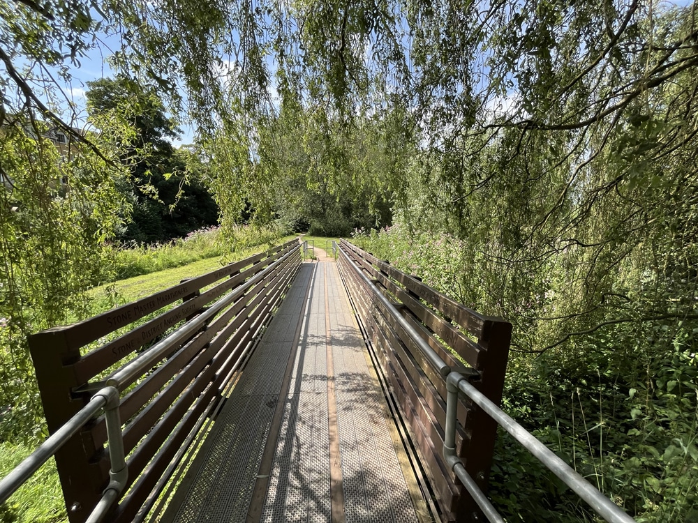 Wooden footbridge with metal railings crossing into Crown Meadow, shaded by low-hanging tree branches and surrounded by lush greenery.