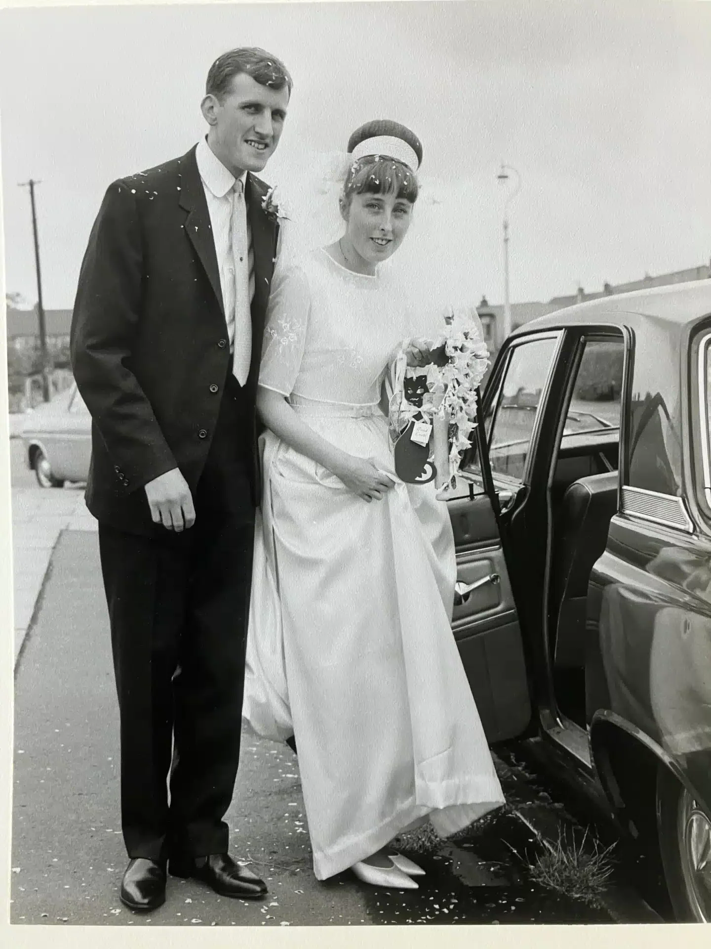 Alan and Anne Taylor on their wedding day in 1965, standing beside a classic car after their Birmingham ceremony
