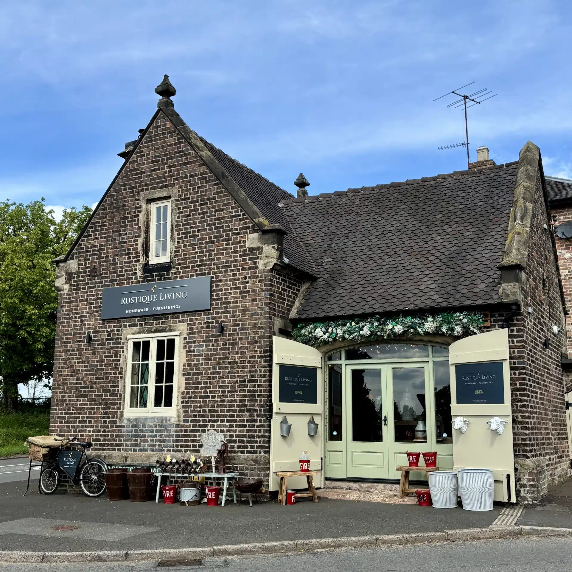 Front of Rustique Living, a homeware store in Sandon, featuring a traditional brick building with pastel green doors, floral decorations, and rustic outdoor displays.