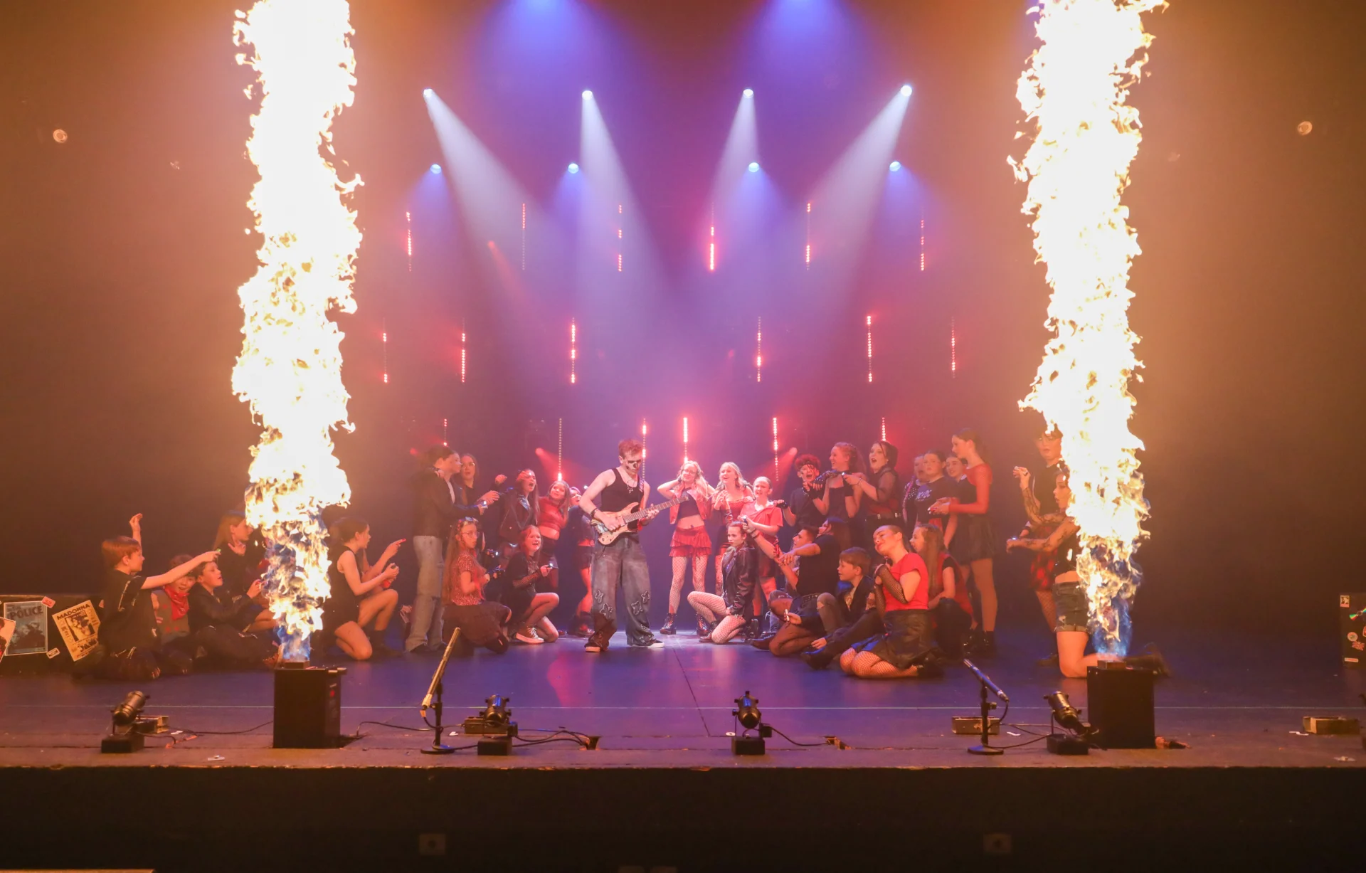 Performers from Rooftop Studios on stage at The Regent Theatre, gathered around a guitarist with dramatic fire effects and stage lighting during their Believe! show finale.