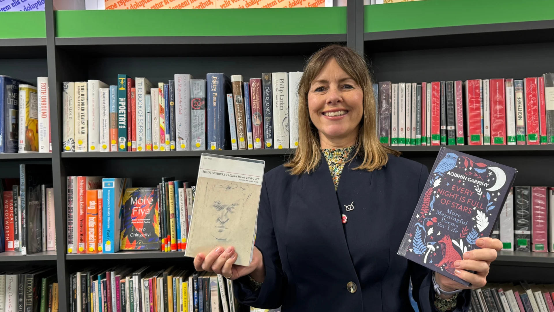 Hayley Coles holding two poetry books in front of shelves at a Staffordshire library.