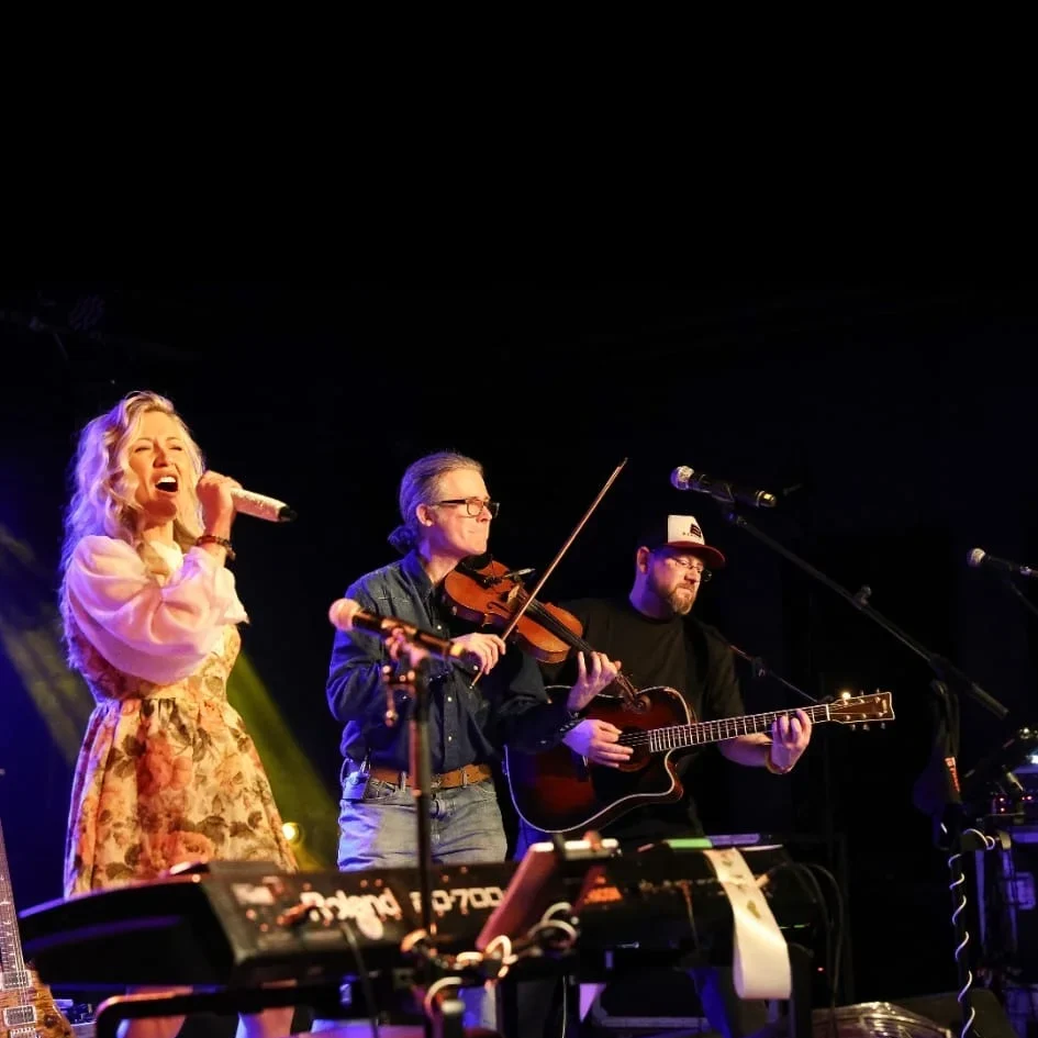 Laura Aston singing on stage with two musicians playing violin and guitar.