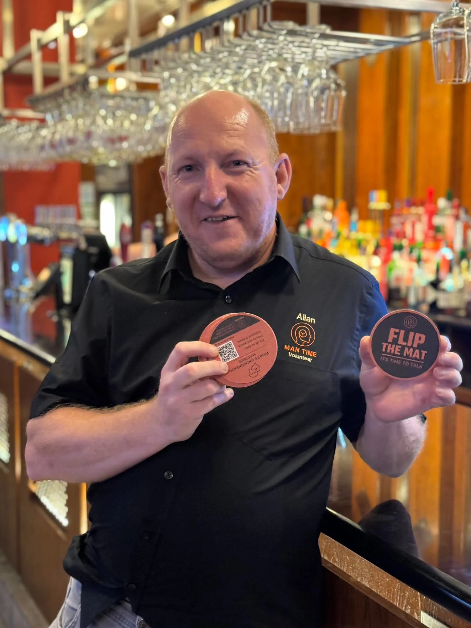 Man Time volunteer, Alan, holding two Flip the Mat campaign beer mats inside The Poste of Stone.