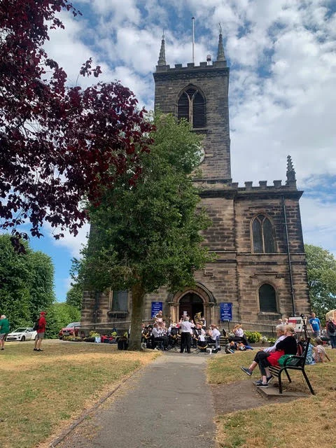 Florence Brass performing outside St Michael and St Wulfad Church in Stone, with people sitting on the church grounds watching.