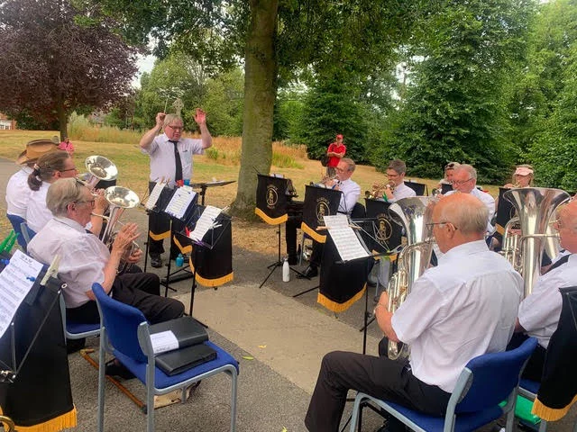 Members of Florence Brass performing outdoors in Stone, with the musical director conducting the band beneath a tree.