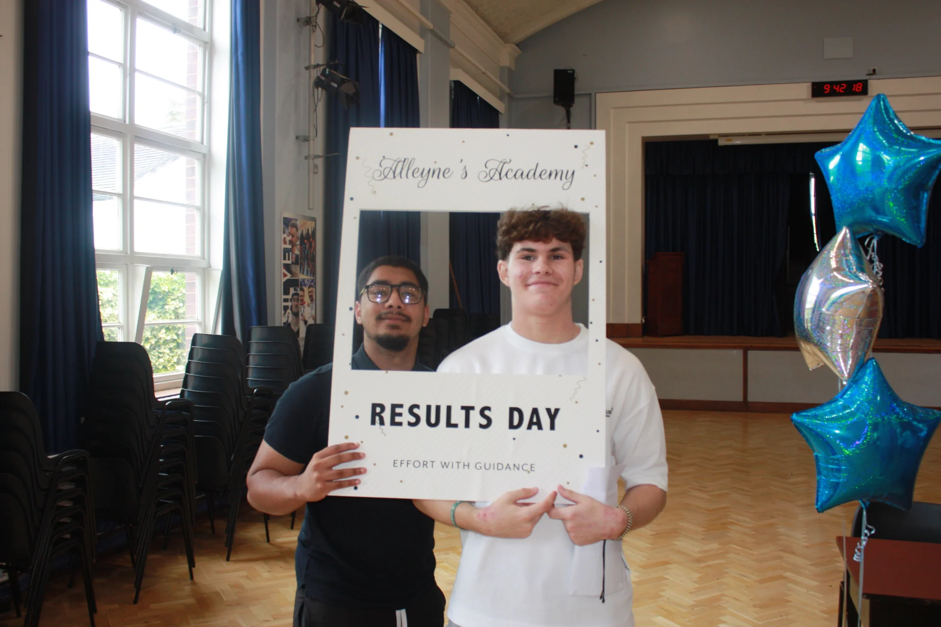 Two Alleyne’s Academy students pose with a Results Day frame and blue and silver balloons inside the school hall in Stone.