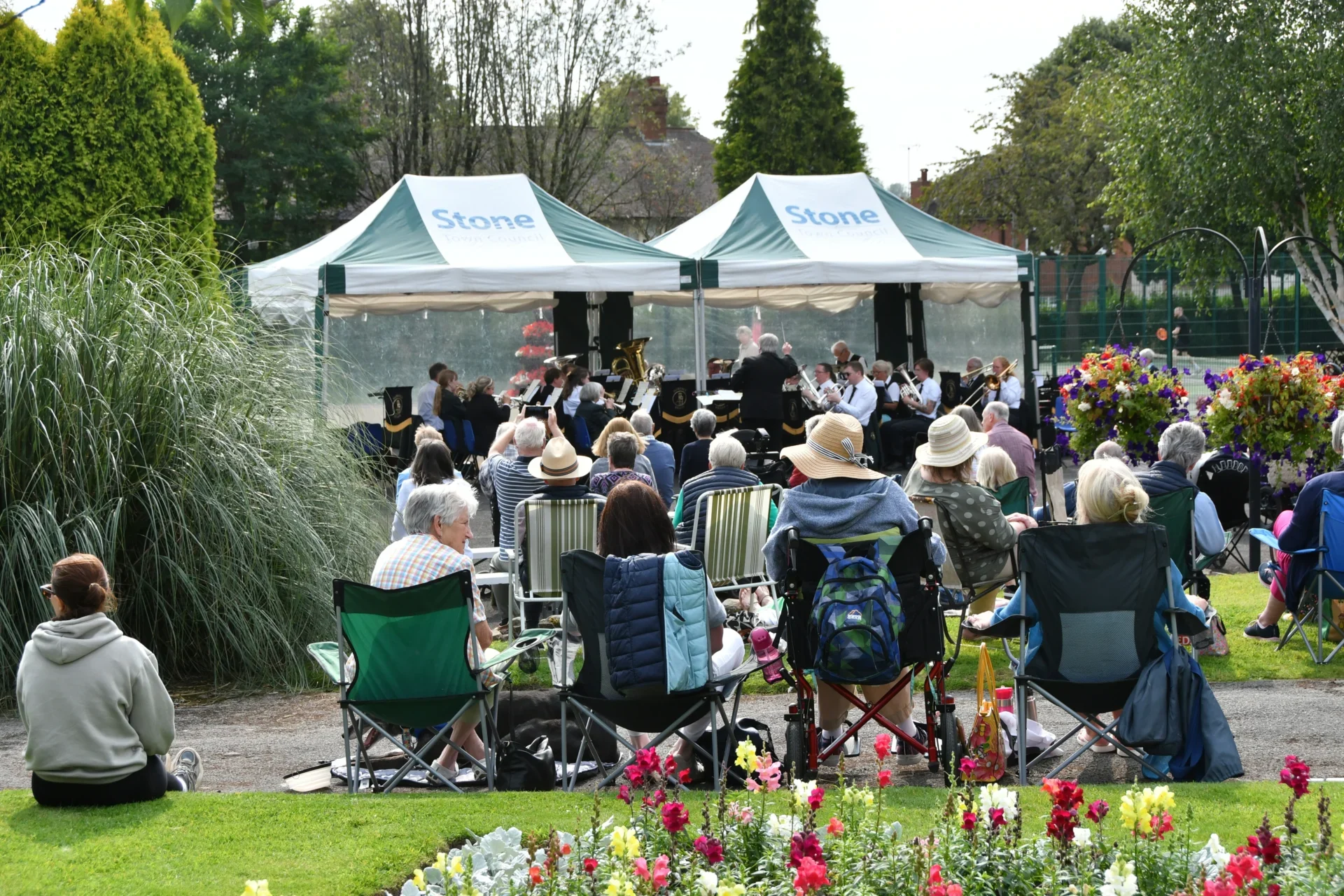 People seated on chairs and picnic blankets watching Florence Brass Band perform beneath Stone Town Council marquees at Stonefield Park in Stone, Staffordshire.