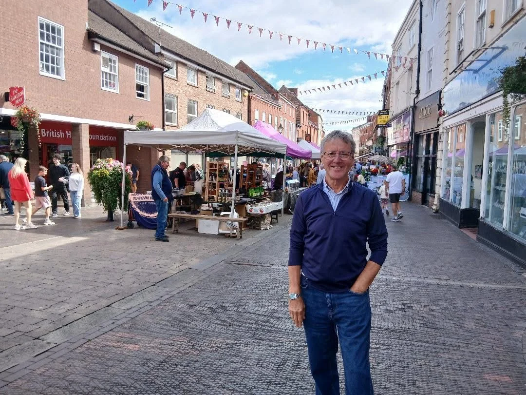 A man standing in Stone High Street while market stalls are operating along the pedestrianised town centre.