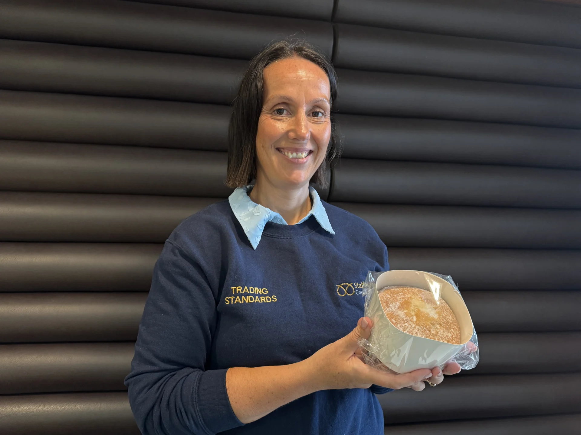 A Staffordshire County Council Trading Standards officer wearing a navy sweatshirt and holding a wrapped cake.