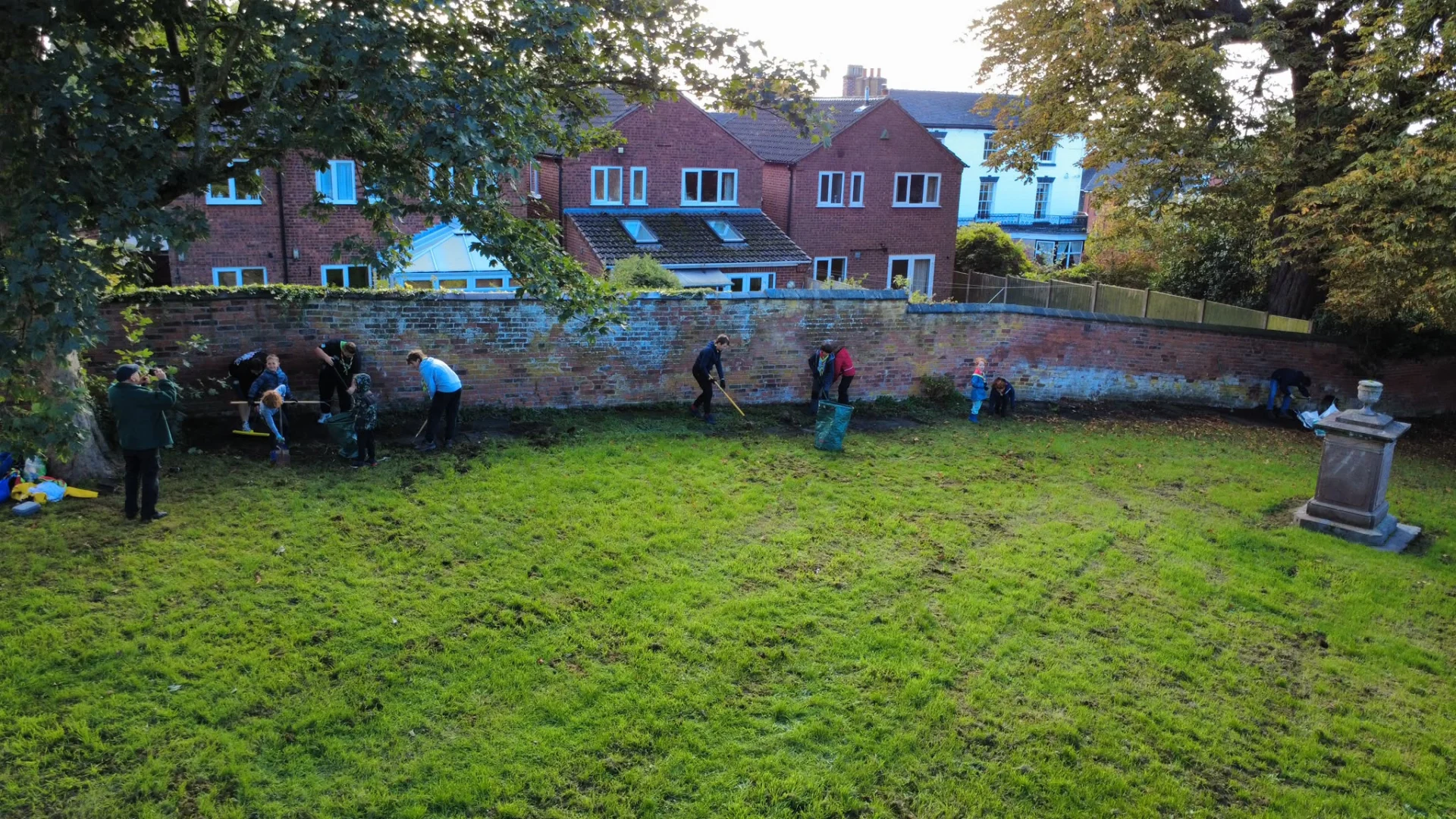 Adults and children clearing vegetation from around headstones beside a brick wall in St Michael and St Wulfad’s churchyard in Stone.