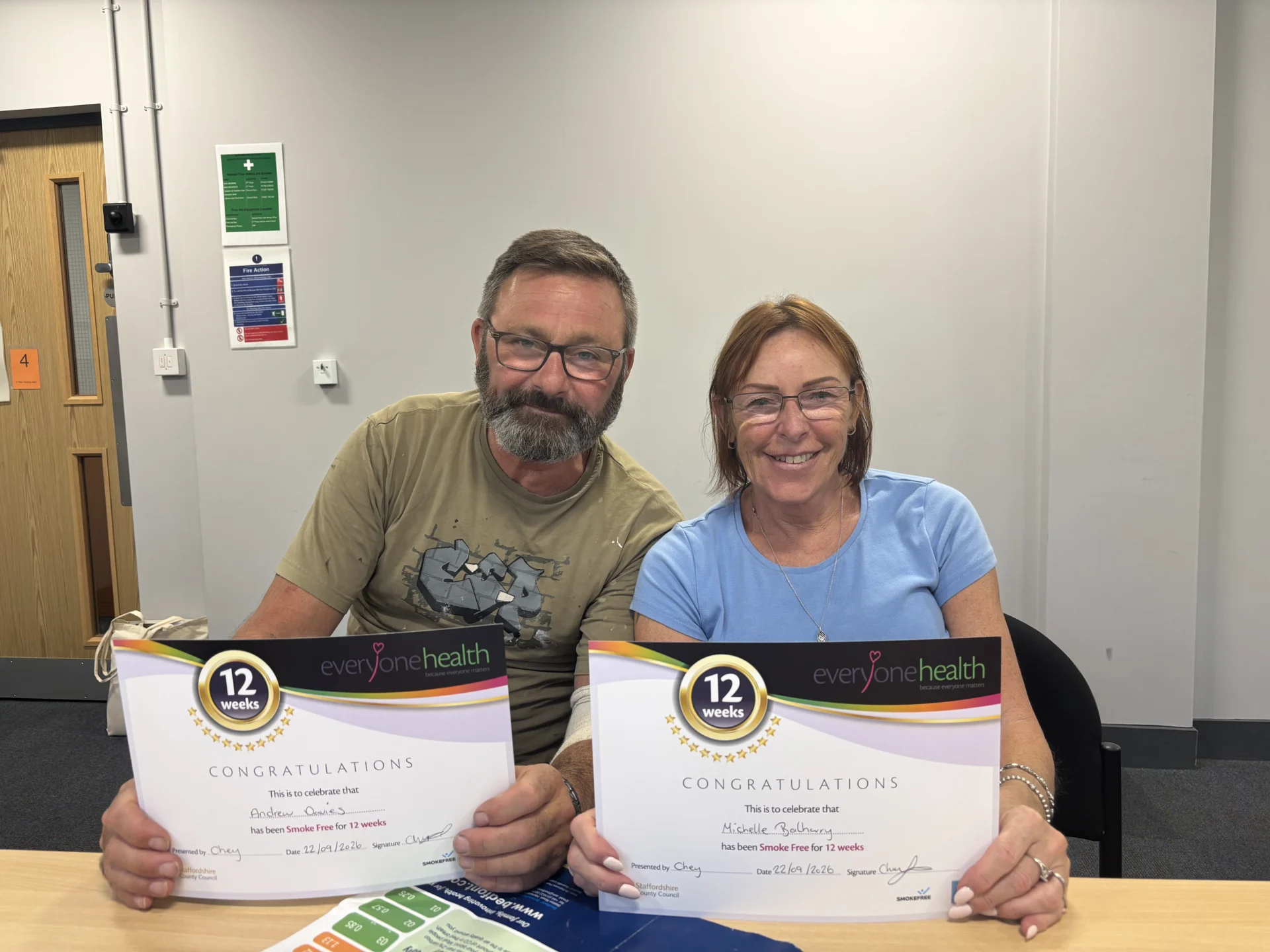 A man and woman sitting at a table holding certificates marking 12 weeks smoke free with Everyone Health Staffordshire.