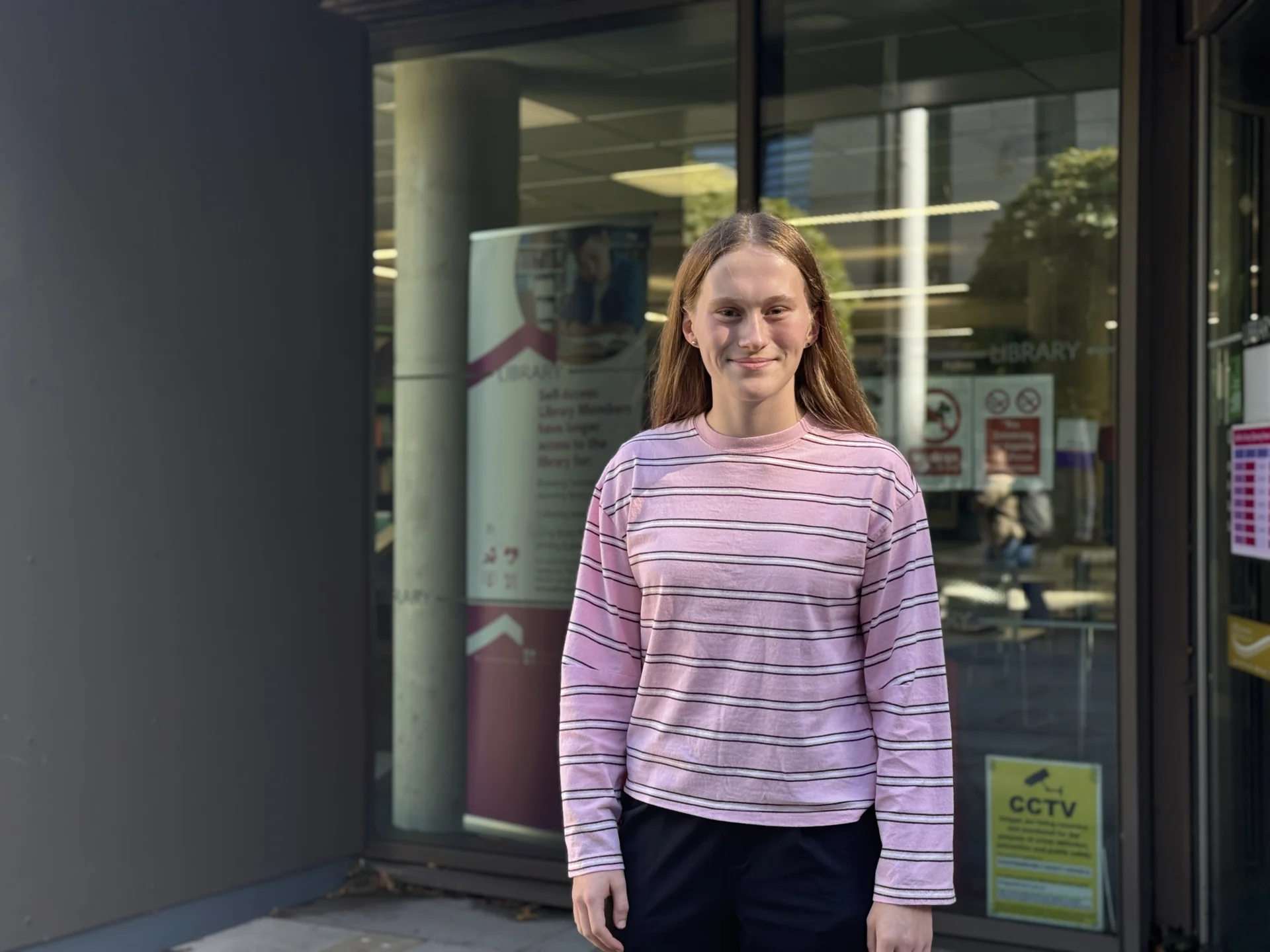 Lola wearing a pink striped top standing outside the entrance to a library.