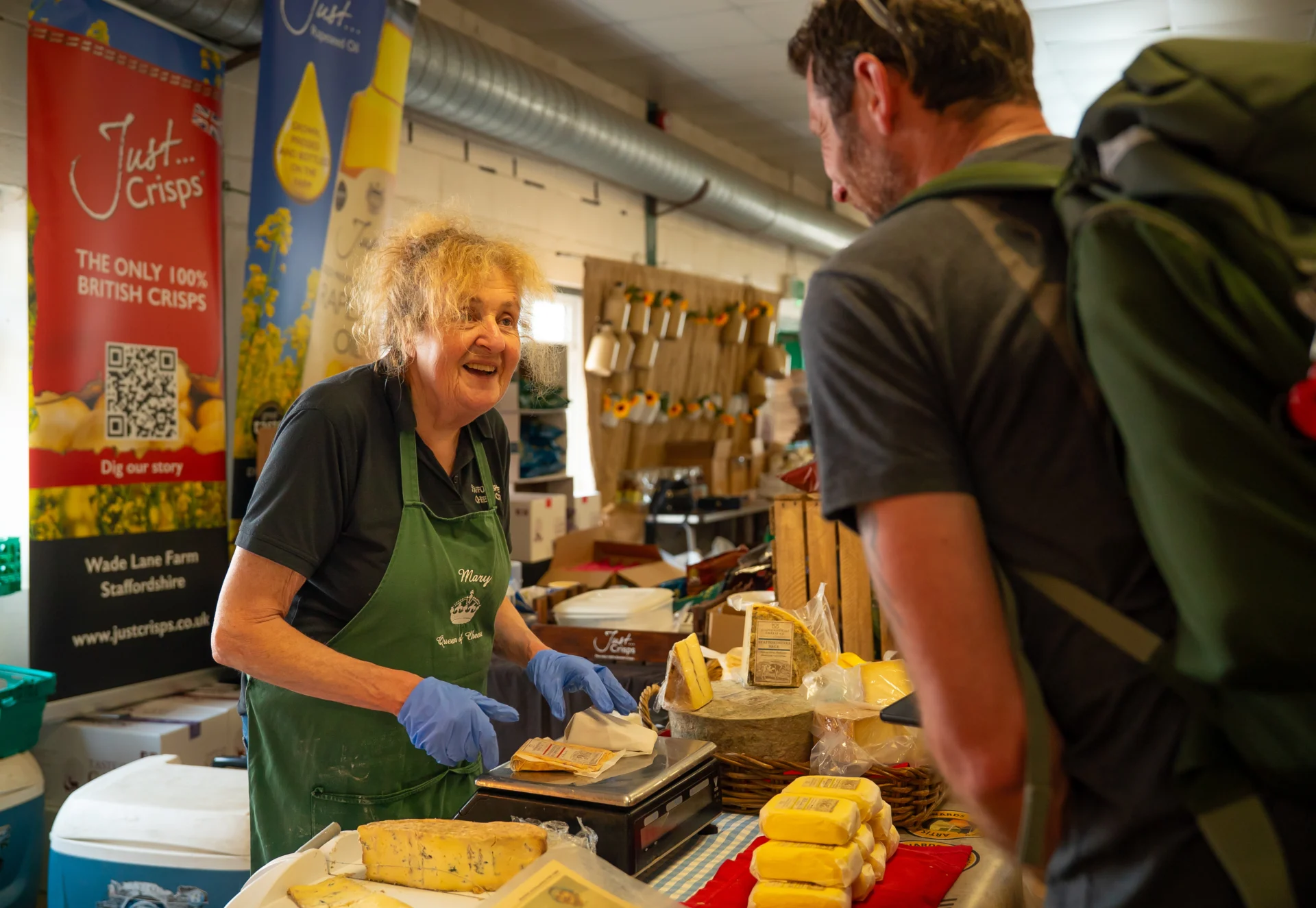 A woman wearing a green apron serves cheese at a food stall while speaking to a customer, with cheeses and other local produce on display.