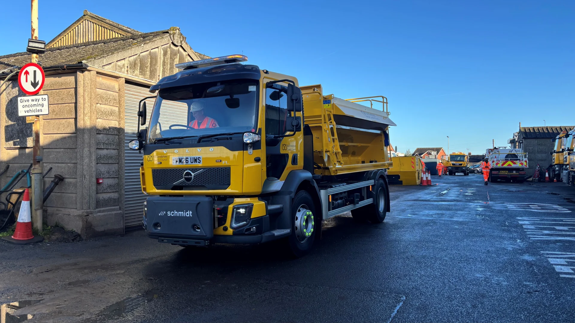 A yellow Staffordshire County Council gritter leaving a highways depot yard, with other council vehicles and workers in the background.