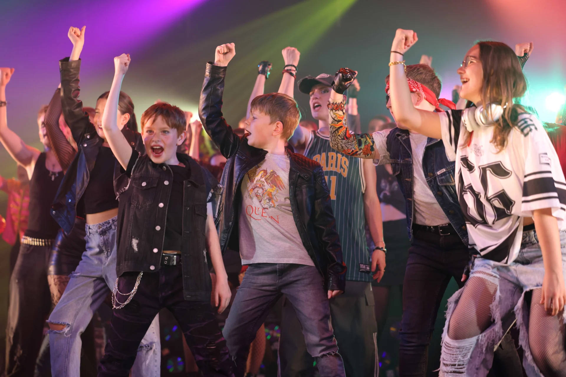 Young Rooftop Studios performers raising their arms on stage, wearing rock inspired outfits under colourful stage lighting.