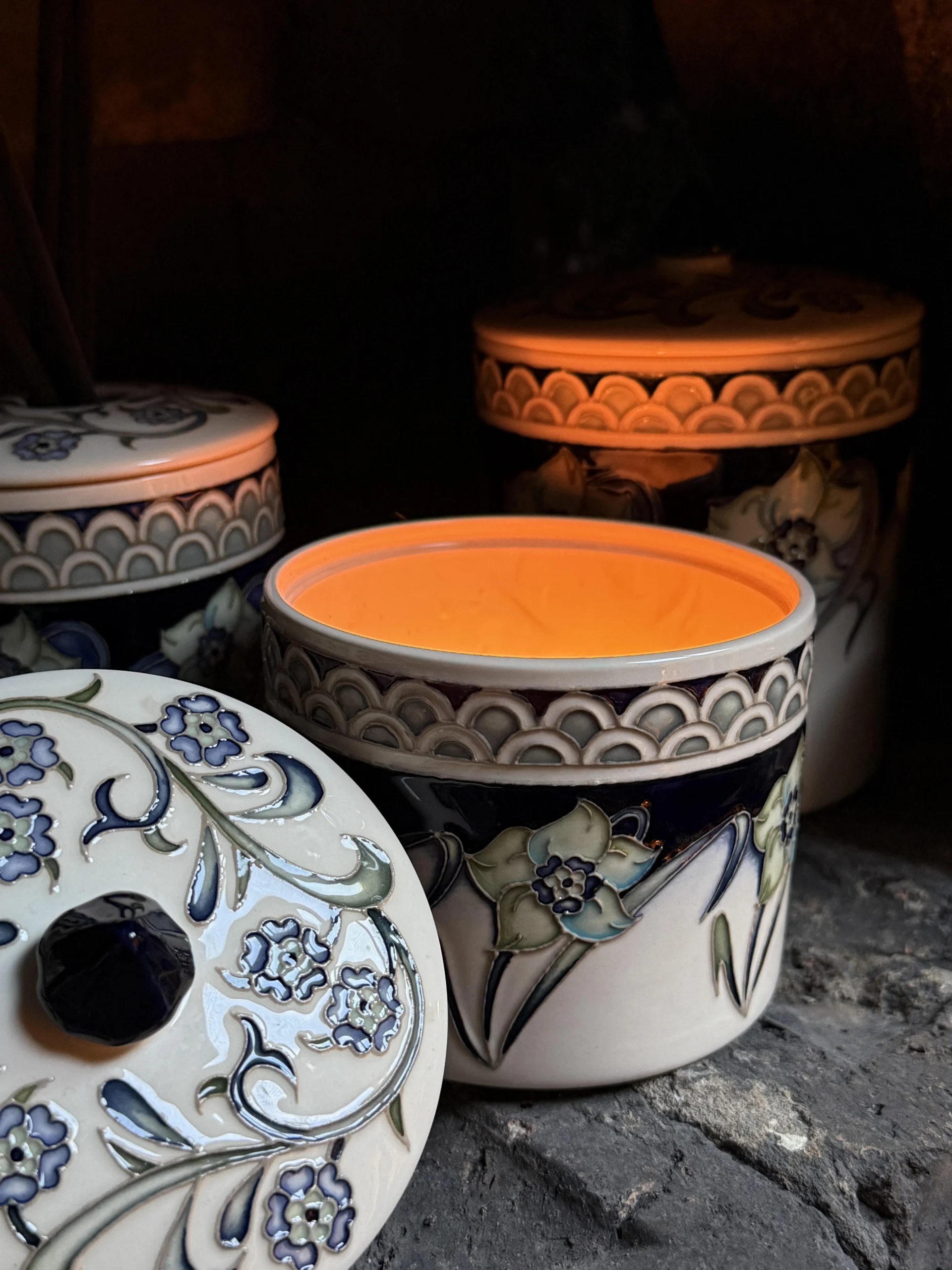 Close view of a lit Moorcroft ceramic candle jar from The Wandering Bee collaboration, with its decorated lid and other matching pieces behind it.